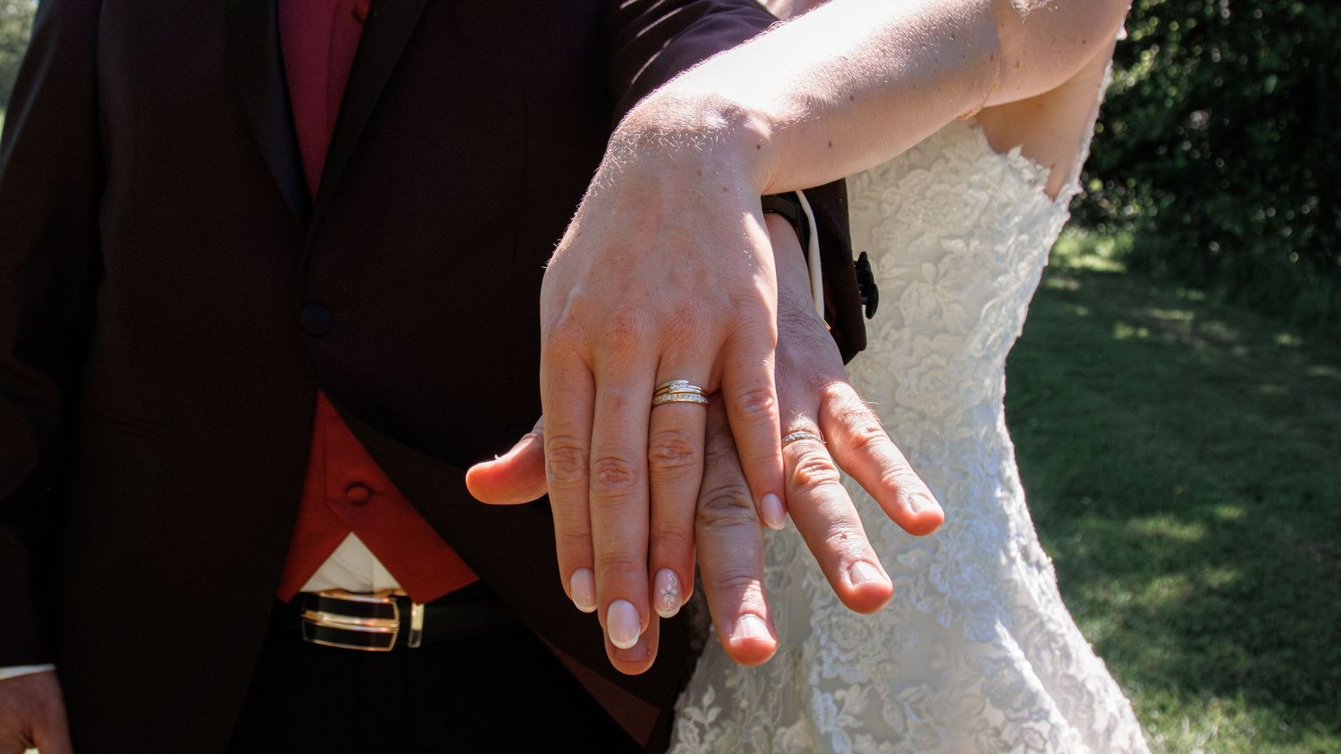 Newlywed couple displaying wedding rings on their hands, bride in lace gown and groom in dark suit with red vest.