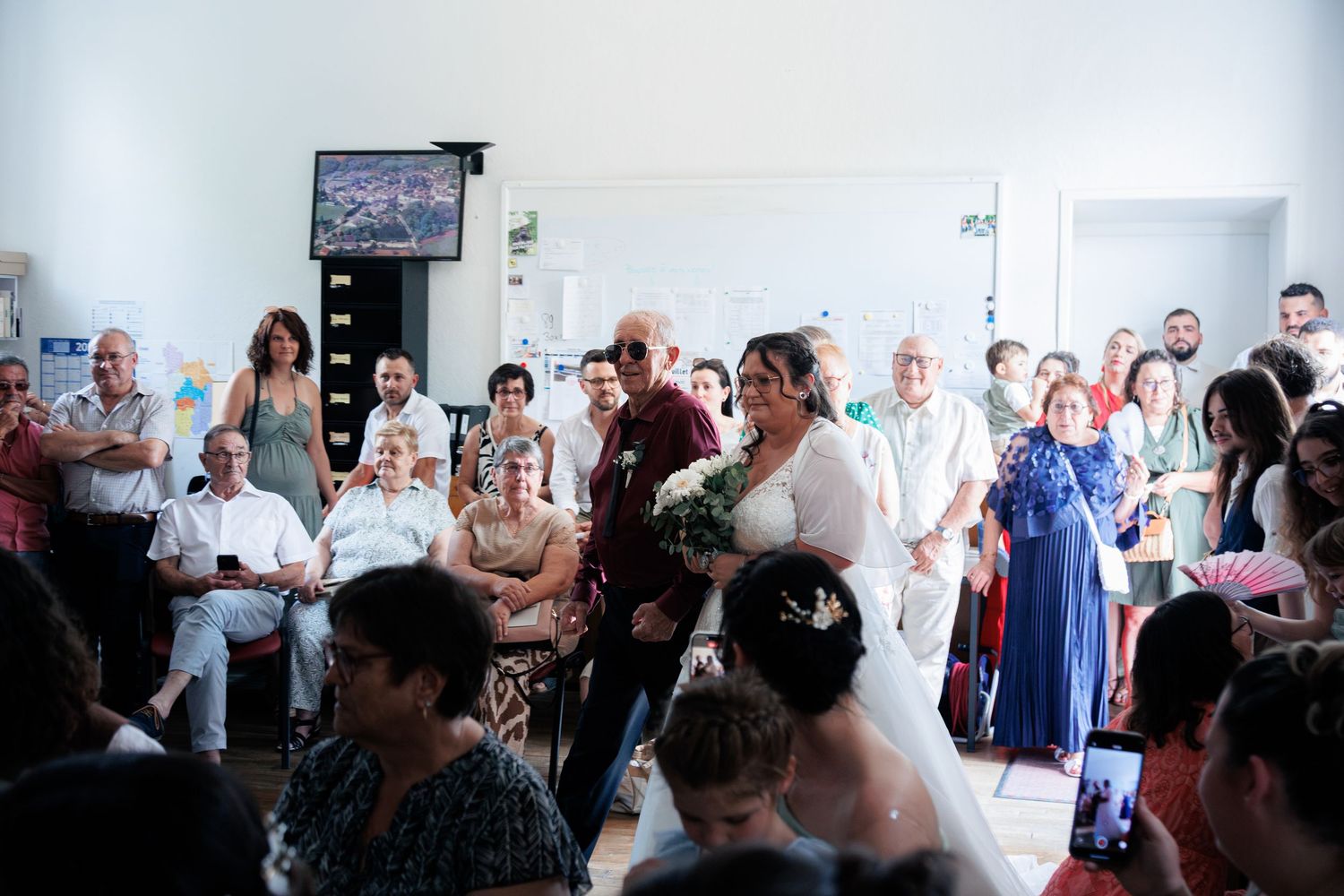 Bride in white gown walks down the aisle with escort at a bright indoor wedding ceremony with seated guests.