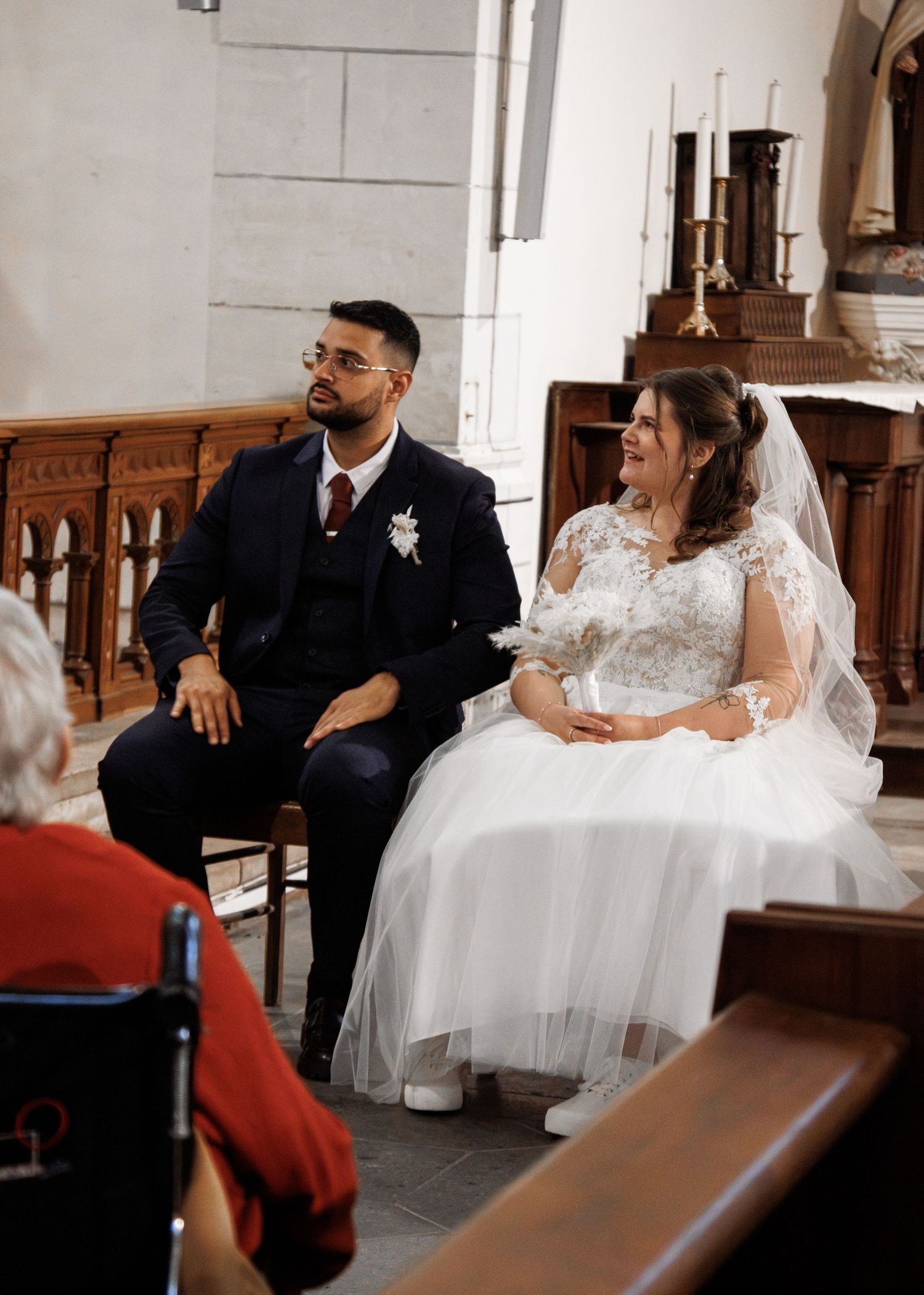 A bride and groom sit together at a church wedding ceremony, with the groom in a navy suit and bride in a white lace gown.
