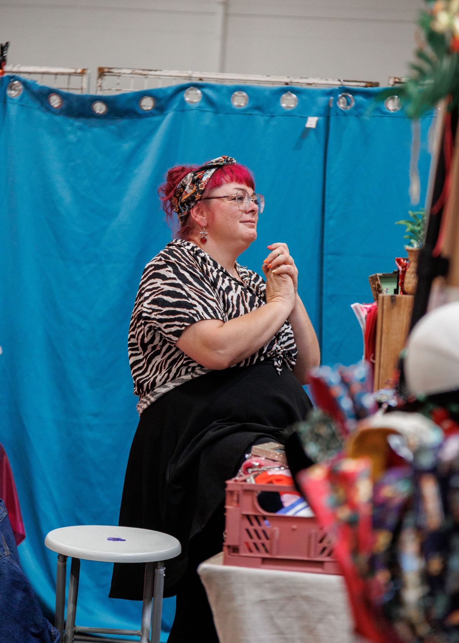 Woman with pink hair and zebra print top sits at a vendor booth with blue backdrop at an indoor market.
