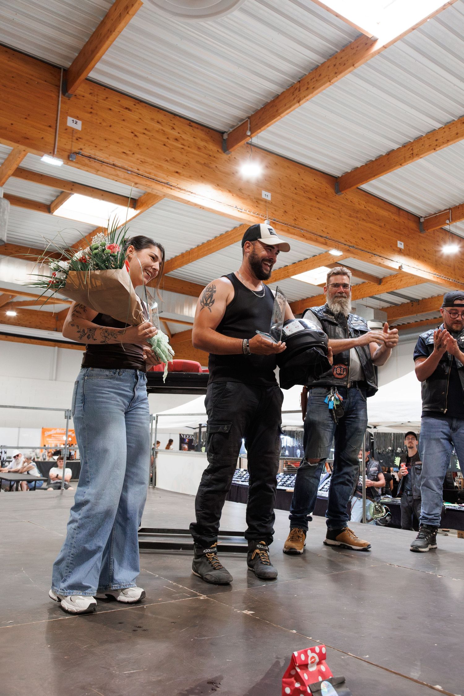 Man presenting flowers and gifts to a woman on stage at an indoor motorcycle event while audience applauds.