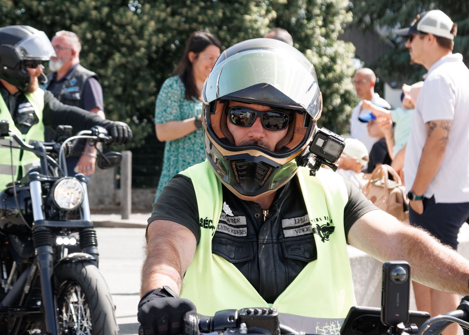 Motorcyclist wearing a white helmet, sunglasses, and hi-vis vest rides a bike at an outdoor event.