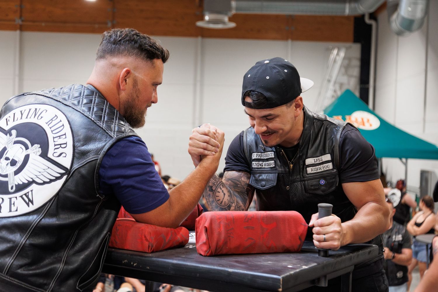 Two men arm wrestling at a biker event, wearing leather vests with patches, competing on a padded table indoors.