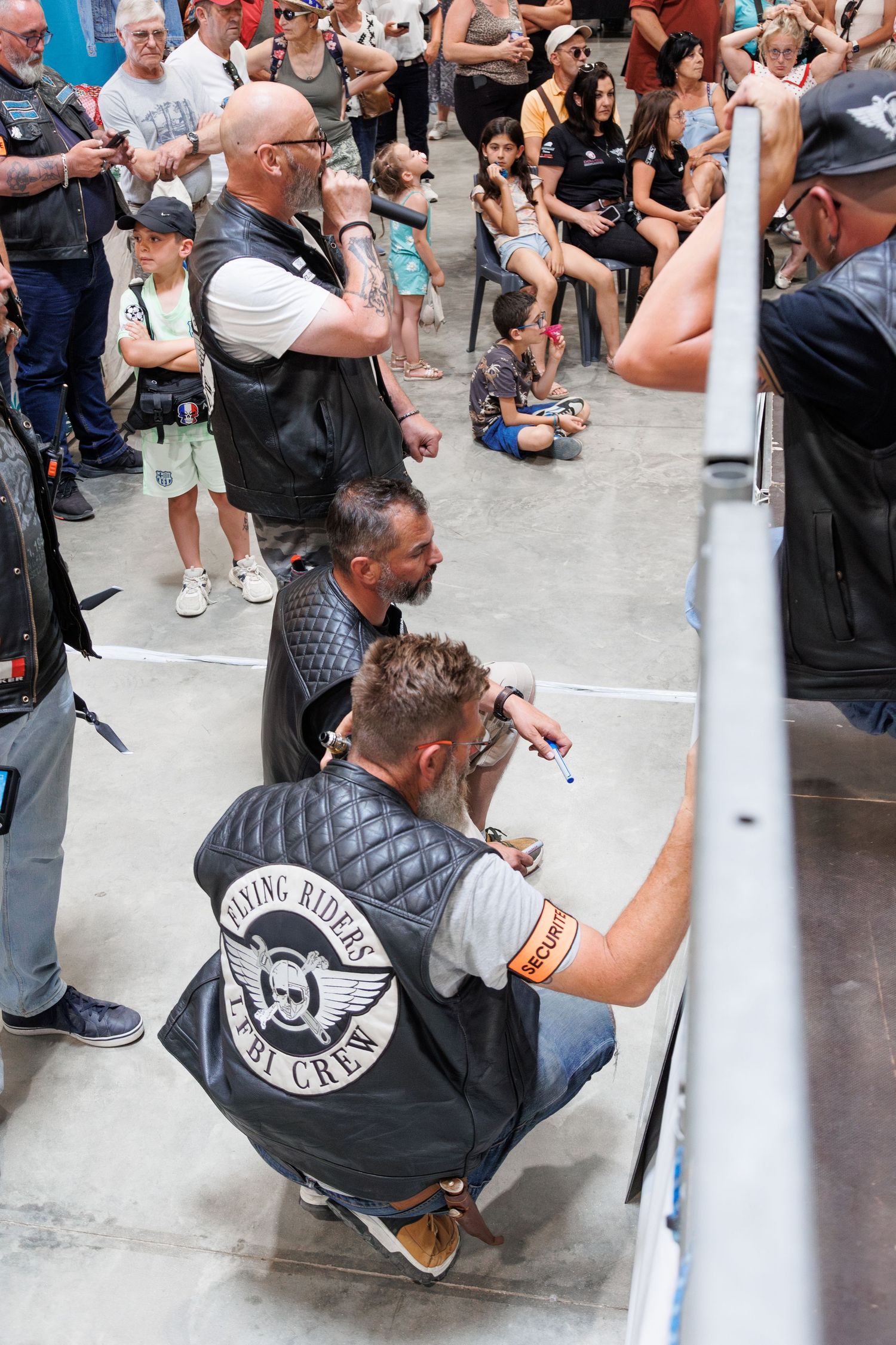 Crowd watches as a man in a Flying Riders Pit Crew vest crouches at a motorsport event indoors.