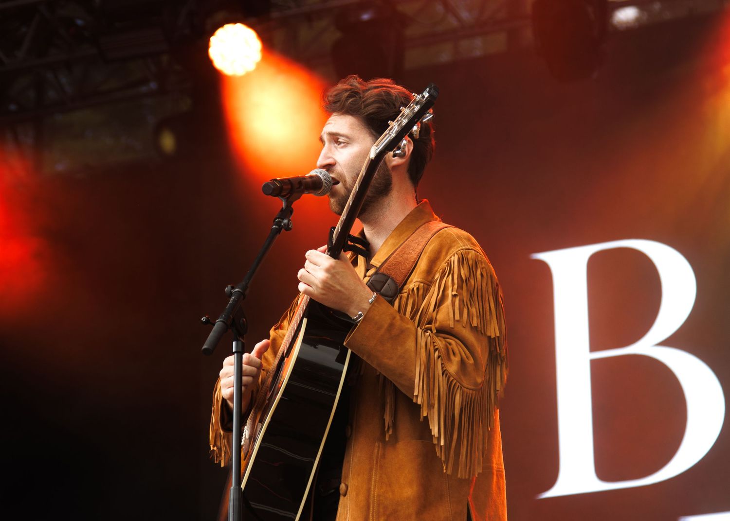A musician performs on stage wearing a brown fringed jacket while playing acoustic guitar with orange lighting and letter B visible.