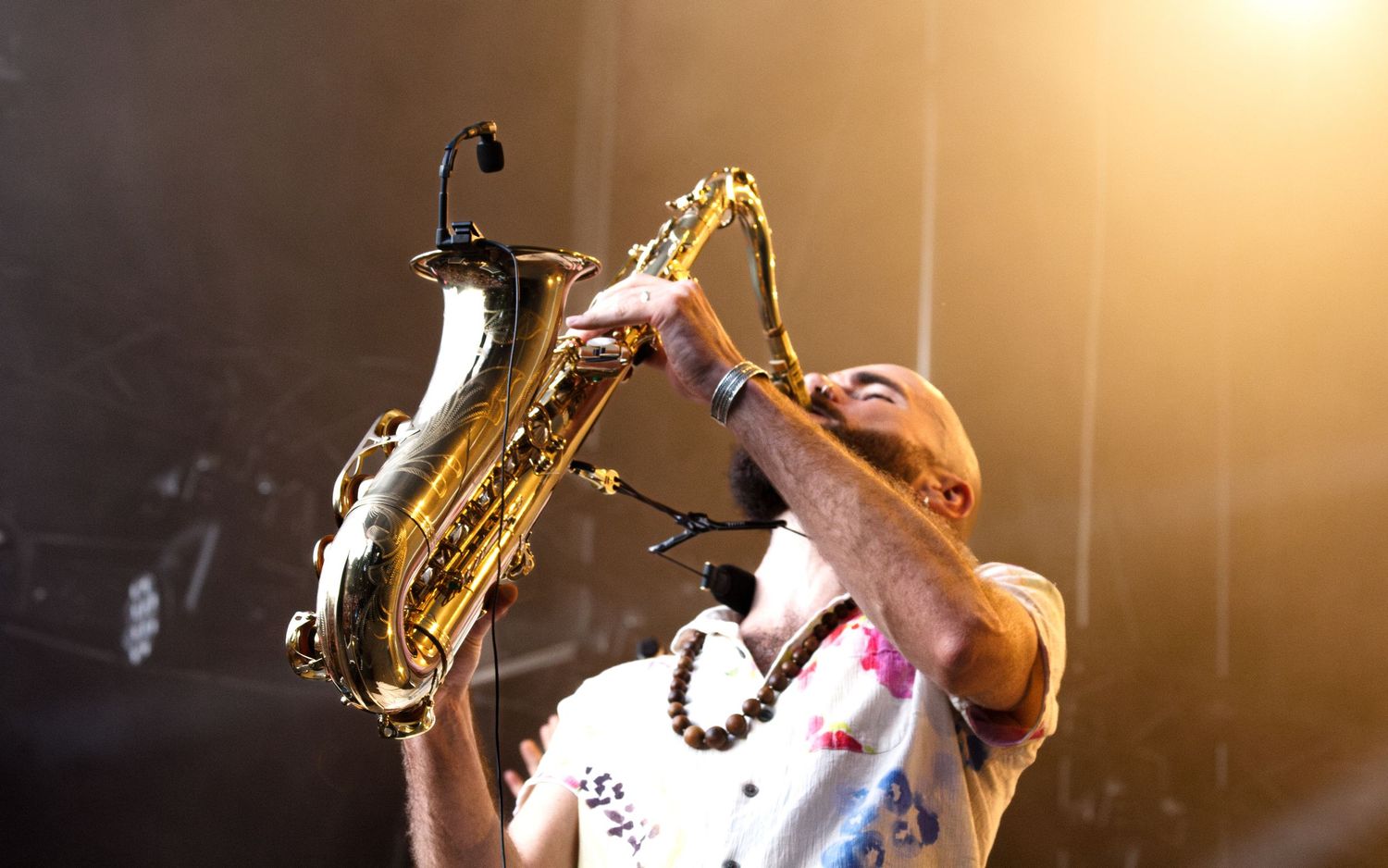 A musician performs on stage with a saxophone illuminated by warm golden stage lighting during a live performance.