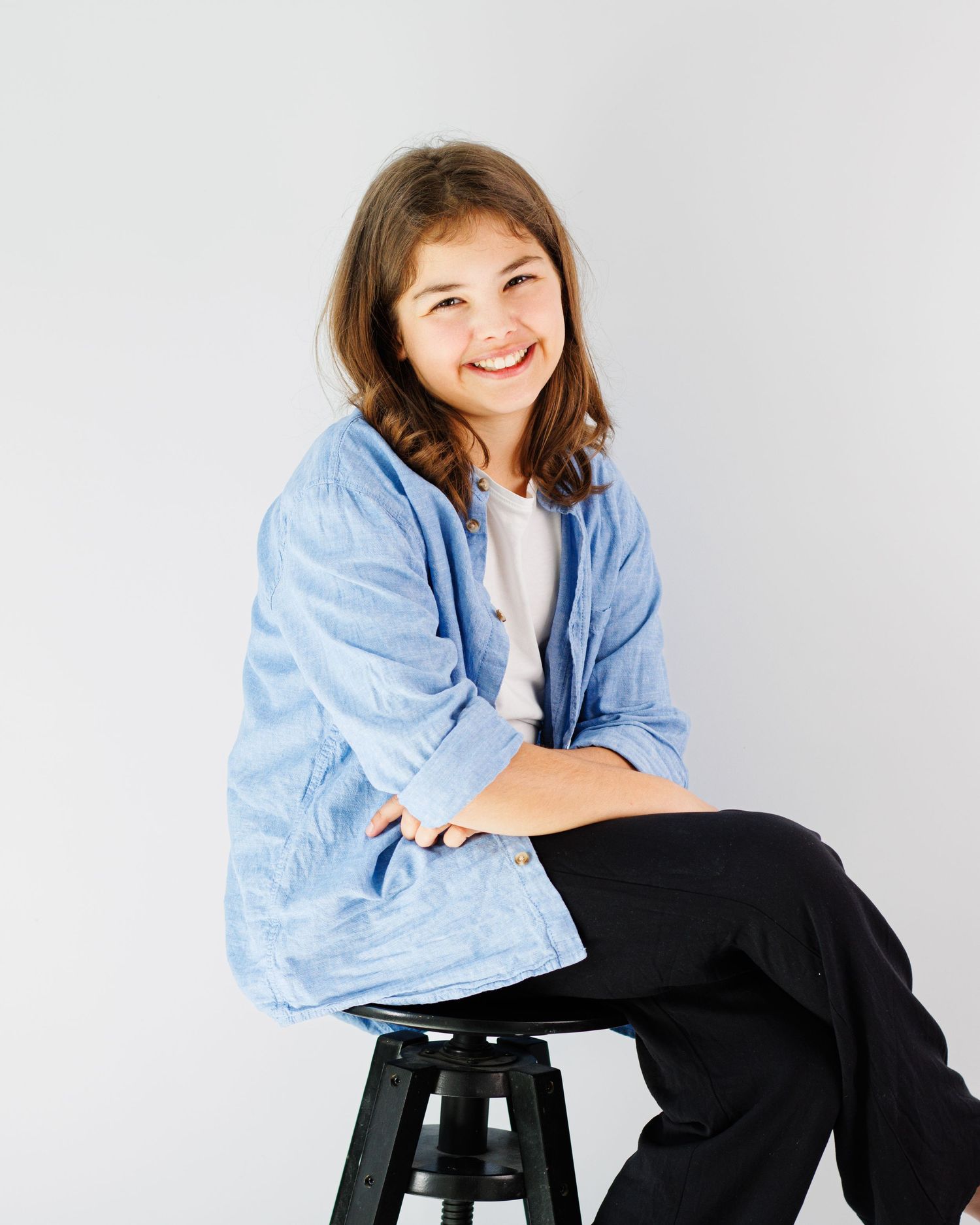 A person in a light blue denim jacket and black pants sits casually on a black stool against a white backdrop.