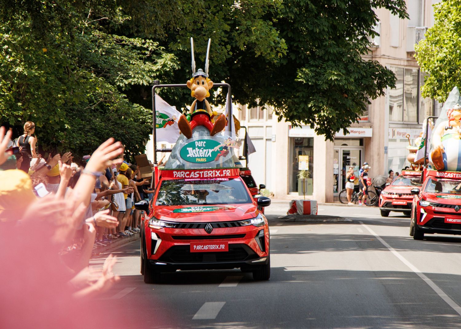 Red promotional vehicle with character mascot on top drives through crowd-lined street.