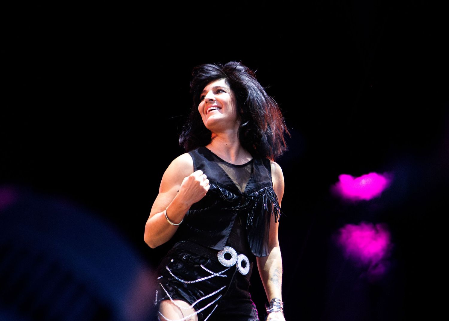 Rock performer with long dark hair plays guitar on stage with pink and blue lighting effects in the background.