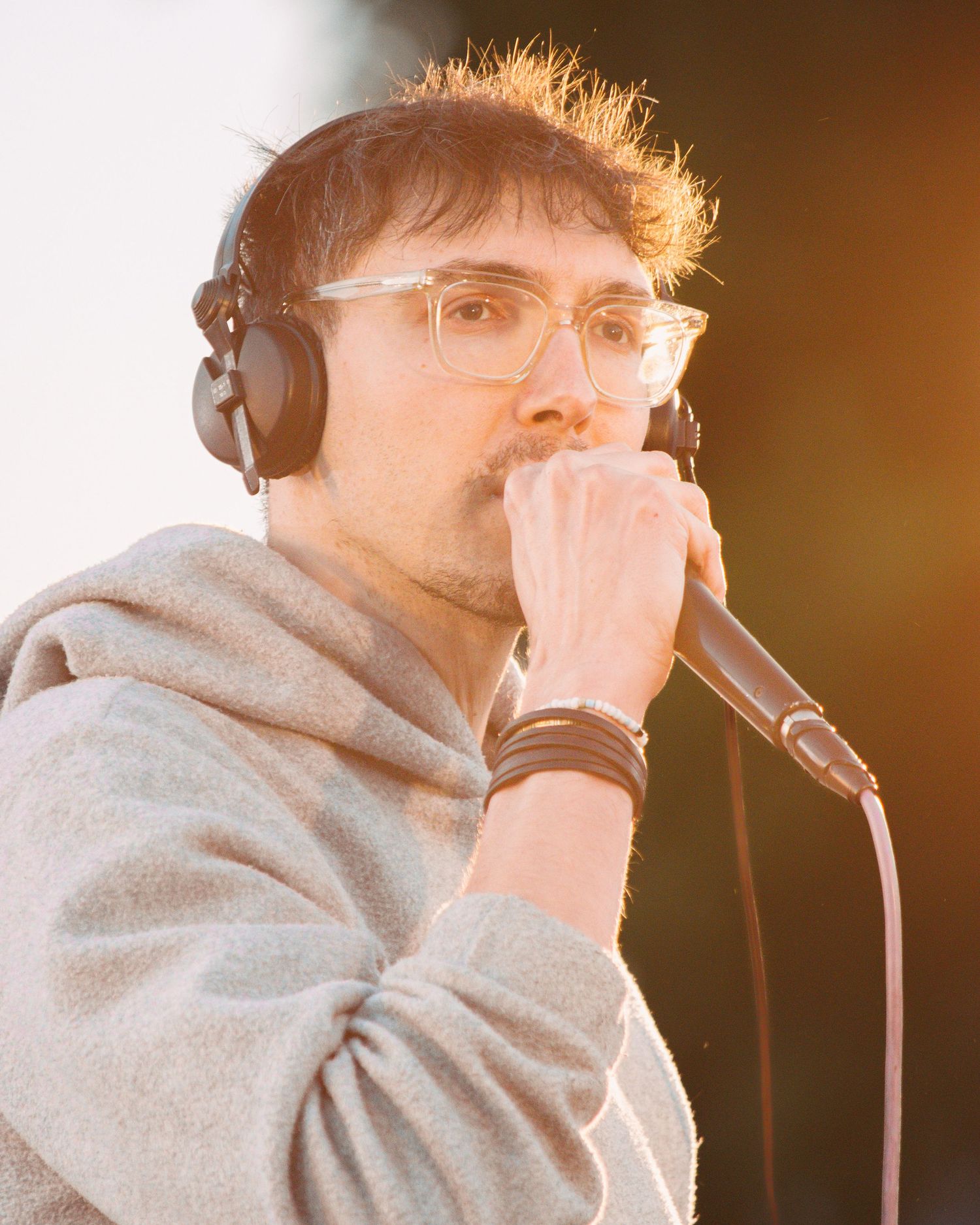 Performer in grey hoodie and headphones singing into microphone during sunset.