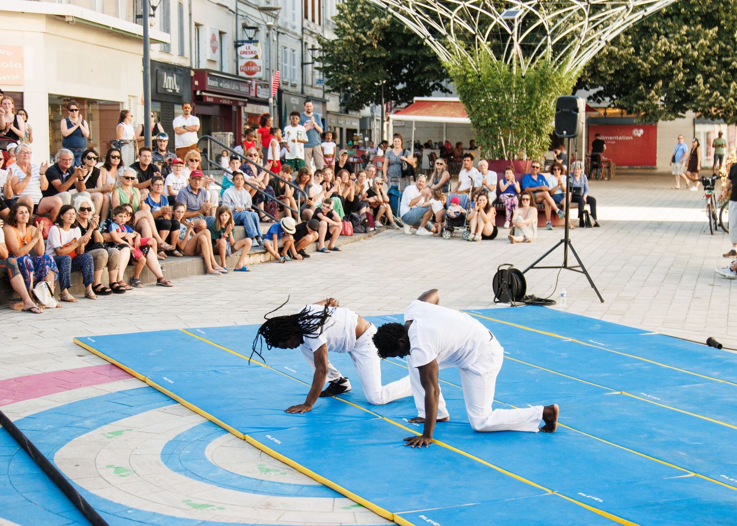 Two performers demonstrate acrobatic moves on a blue performance mat while crowd watches.