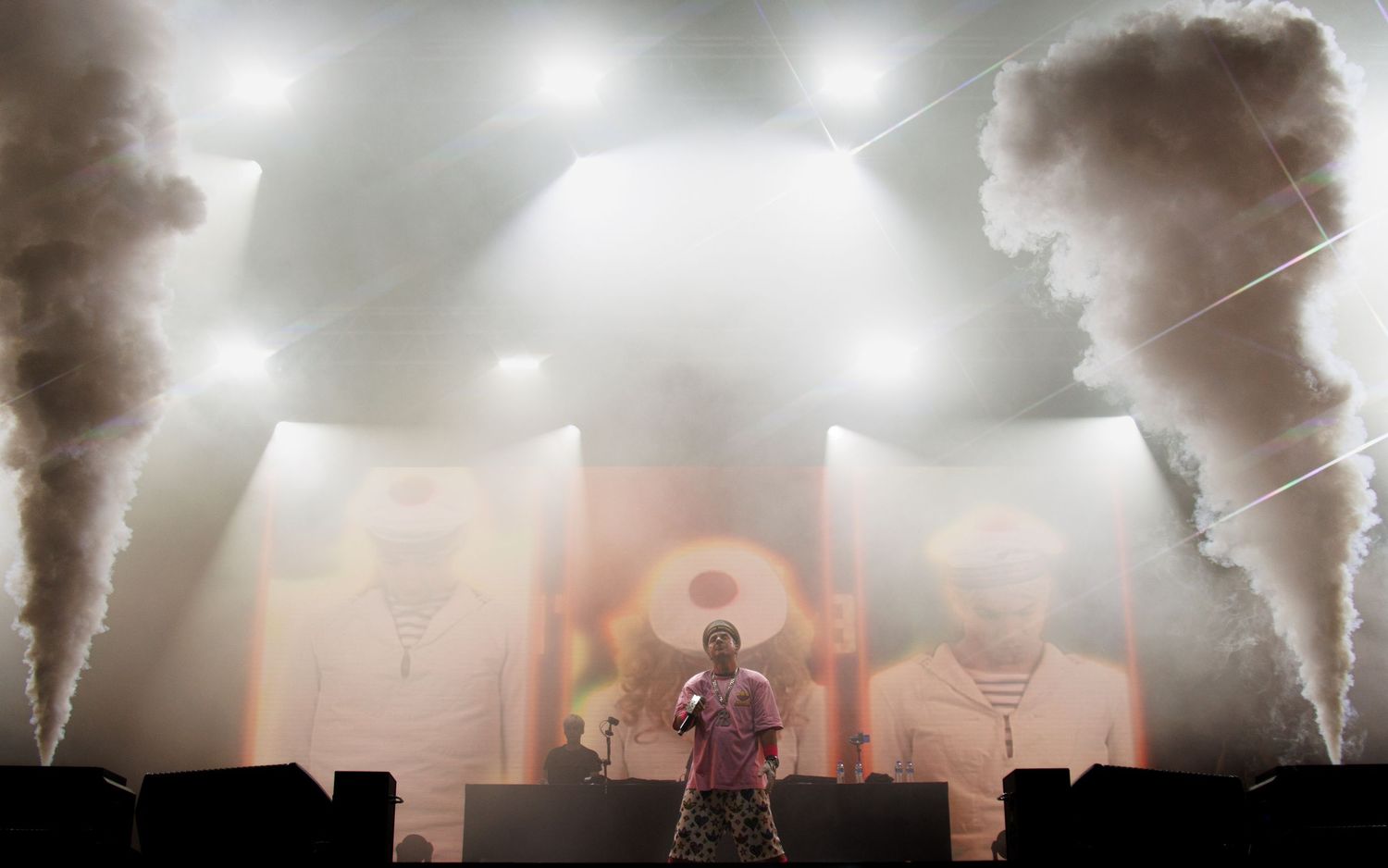 Dramatic concert stage performance with silhouetted performer and multiple smoke machines creating atmospheric effects.