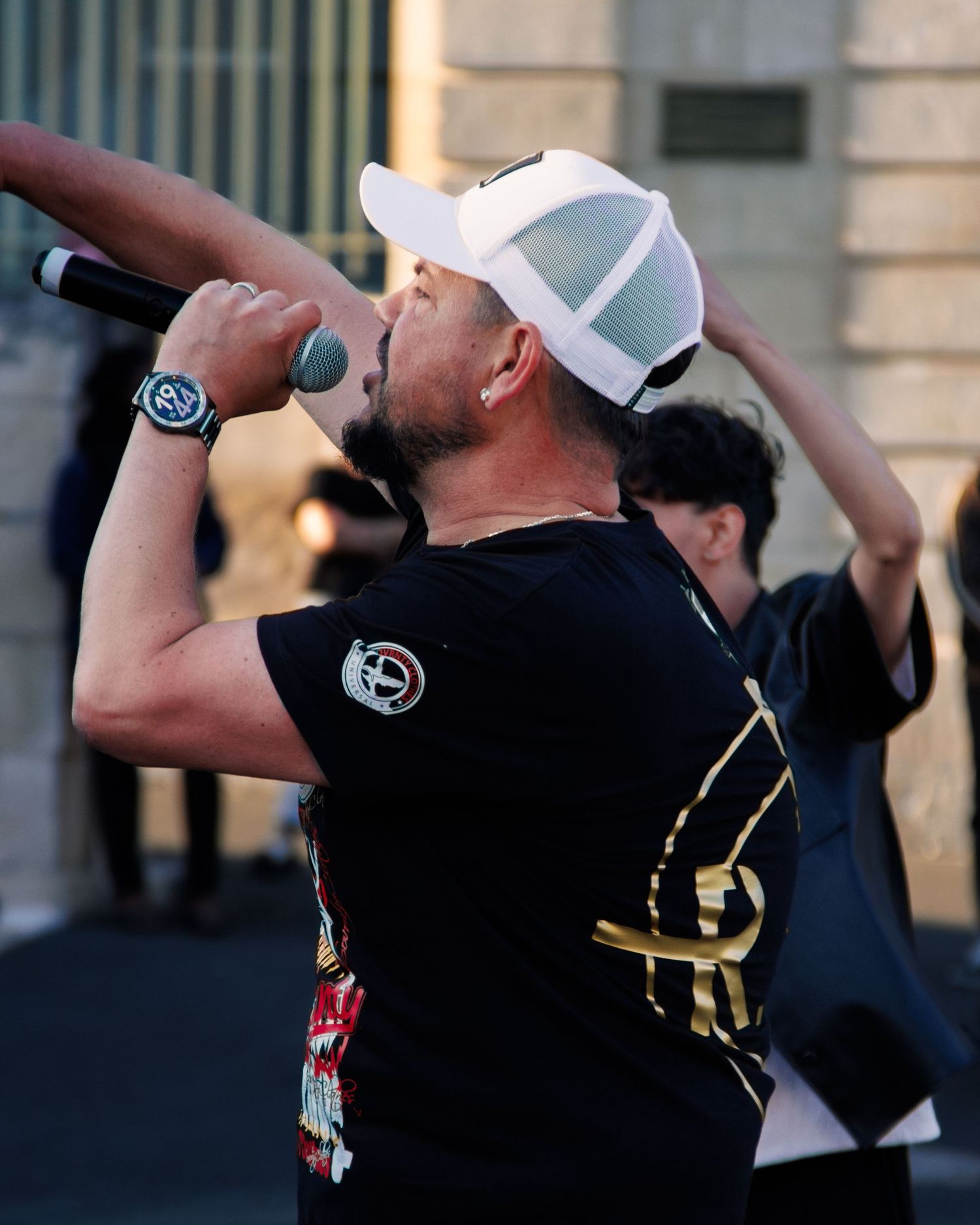 Performer in black shirt and white cap singing into microphone during outdoor event.