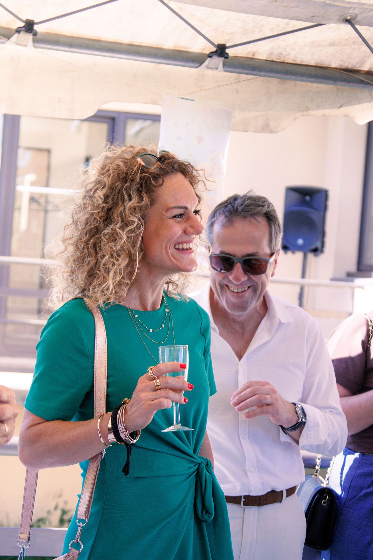 Cheerful woman in teal dress laughing and holding champagne flute at sunny outdoor wedding reception.