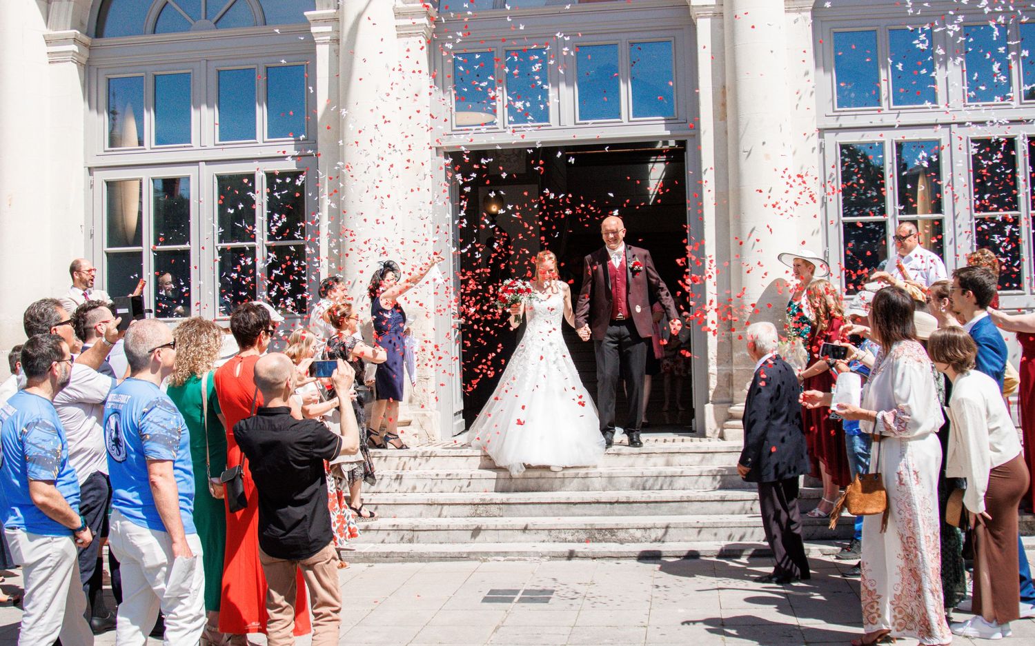 Newlyweds exit grand building through confetti shower surrounded by cheering wedding guests on sunny day.