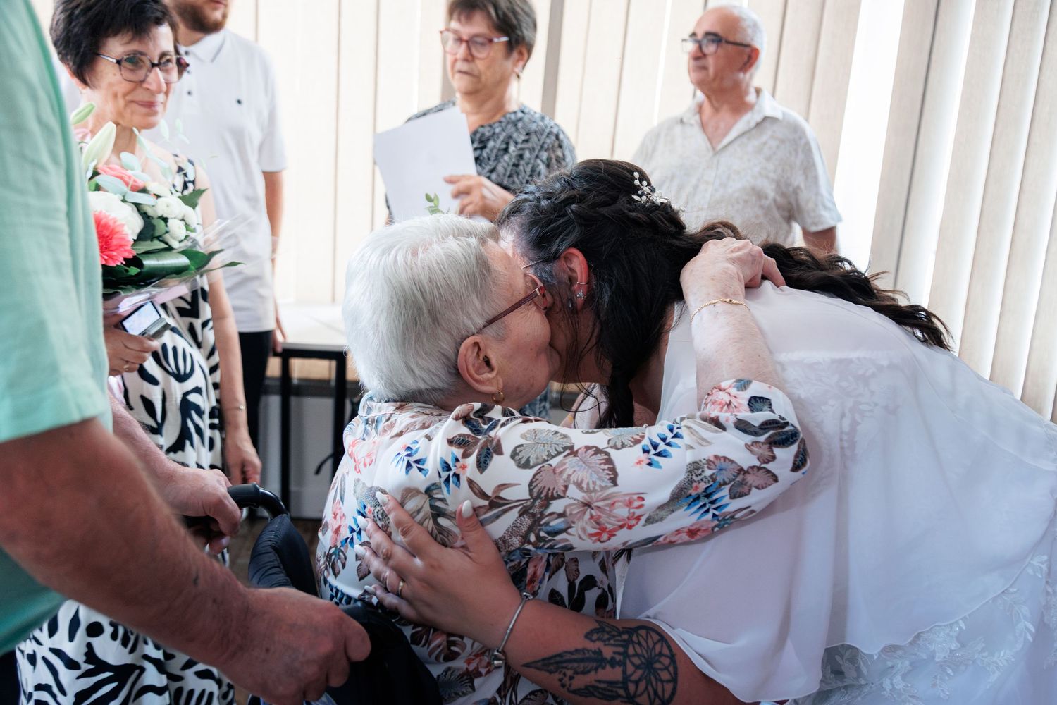 Bride in white gown shares tender embrace with elderly woman in wheelchair at wedding reception.