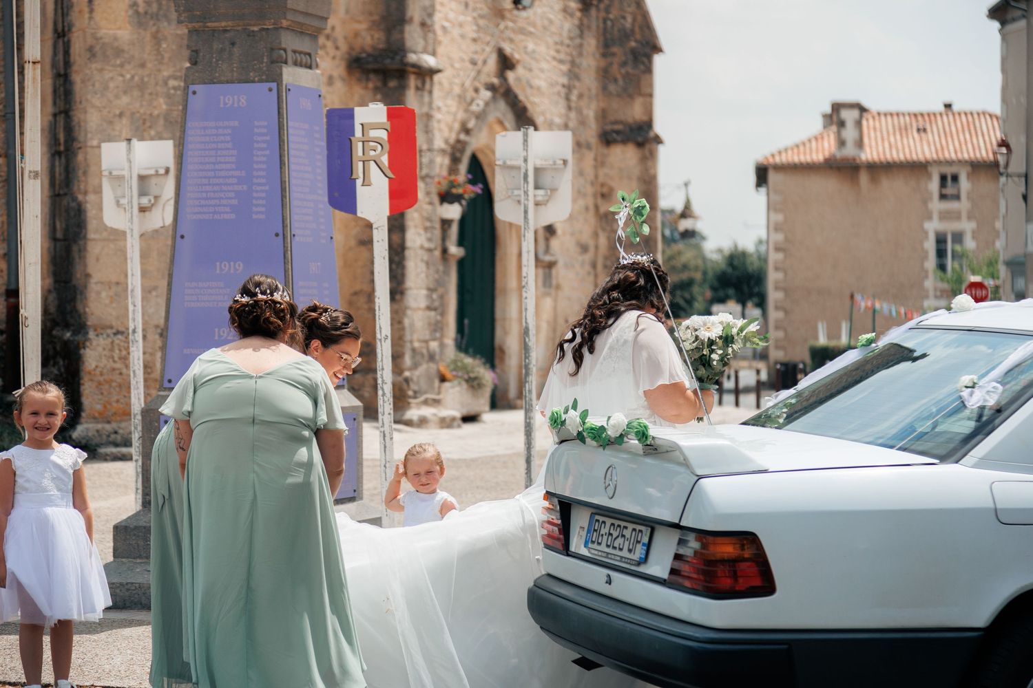 Bride in white gown with bridesmaids in mint green dresses outside a French church with a decorated wedding car.