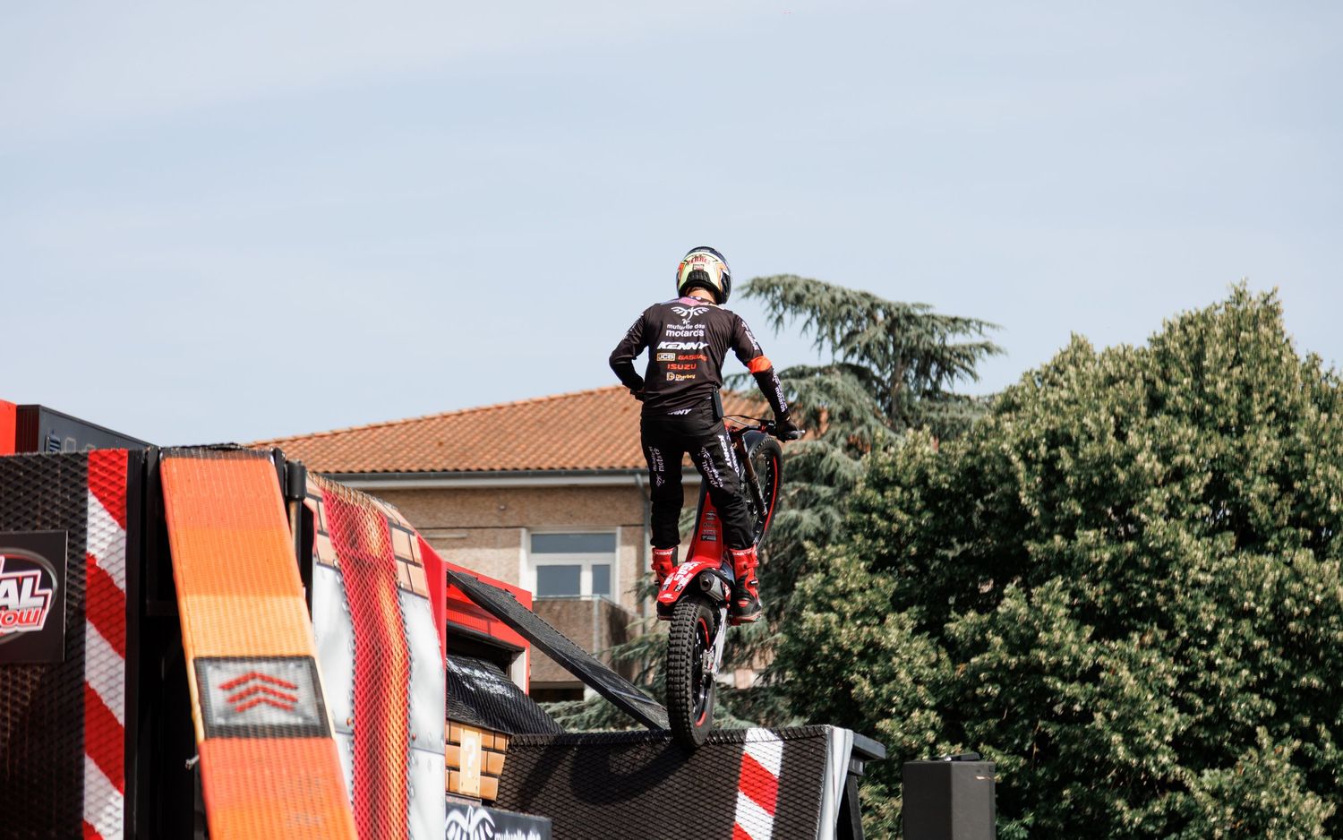 A motocross rider performs a daring stunt on a ramp during an outdoor freestyle competition with trees in the background.