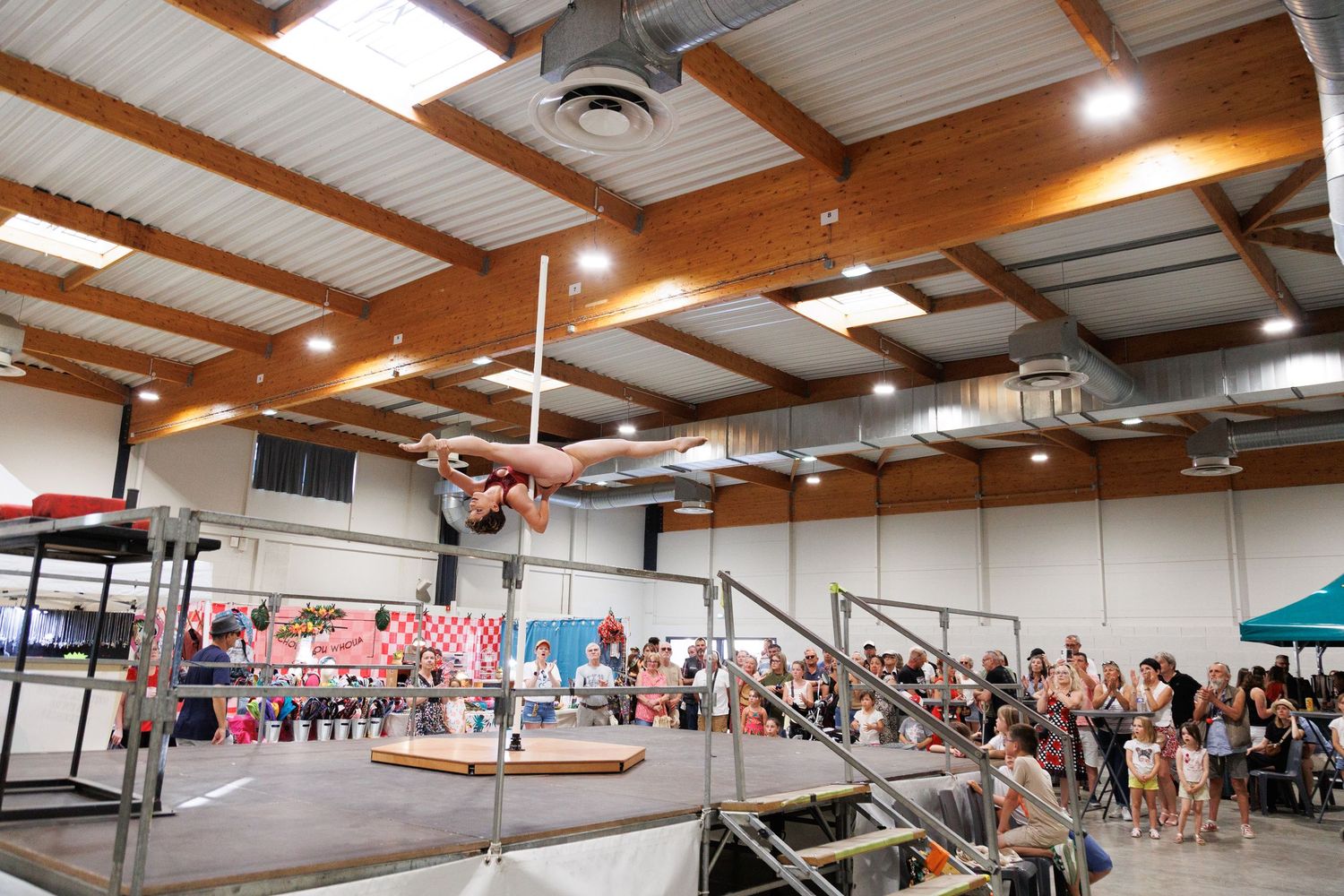 Acrobat performs aerial pole routine inside a large gymnasium with spectators watching during an indoor circus event.