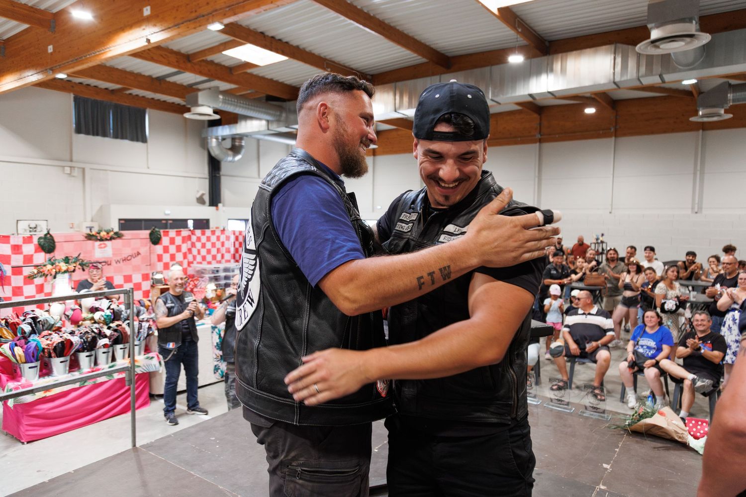 Two men in biker vests share a warm hug at an indoor event with an audience watching in the background.