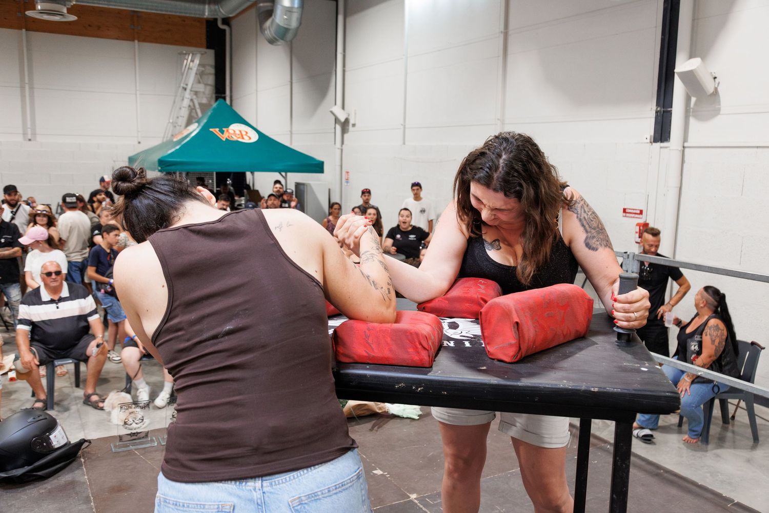Two women competing in an arm wrestling match at an indoor event with spectators watching in the background.