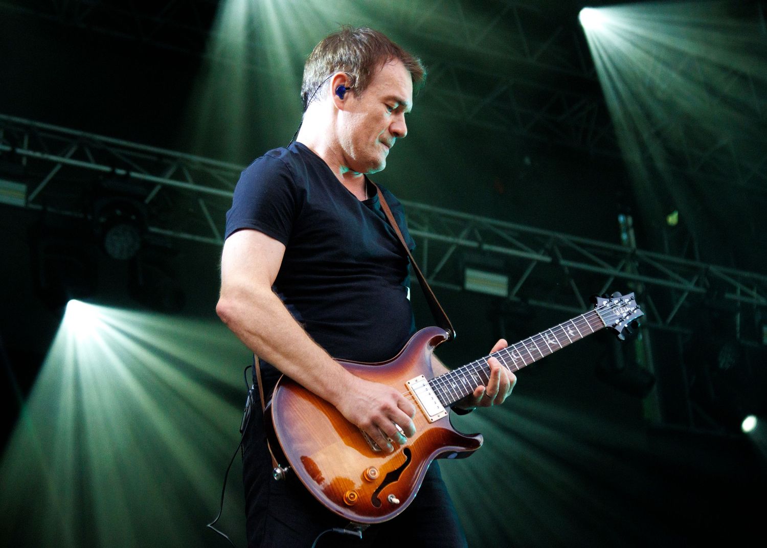 Male guitarist performing on stage under dramatic spotlights, playing a sunburst electric guitar while wearing a black t-shirt.