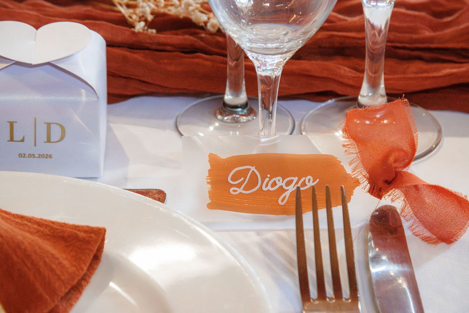 Elegant wedding table setting with orange napkins, gold fork, personalized name card reading Diogo, and crystal glasses.