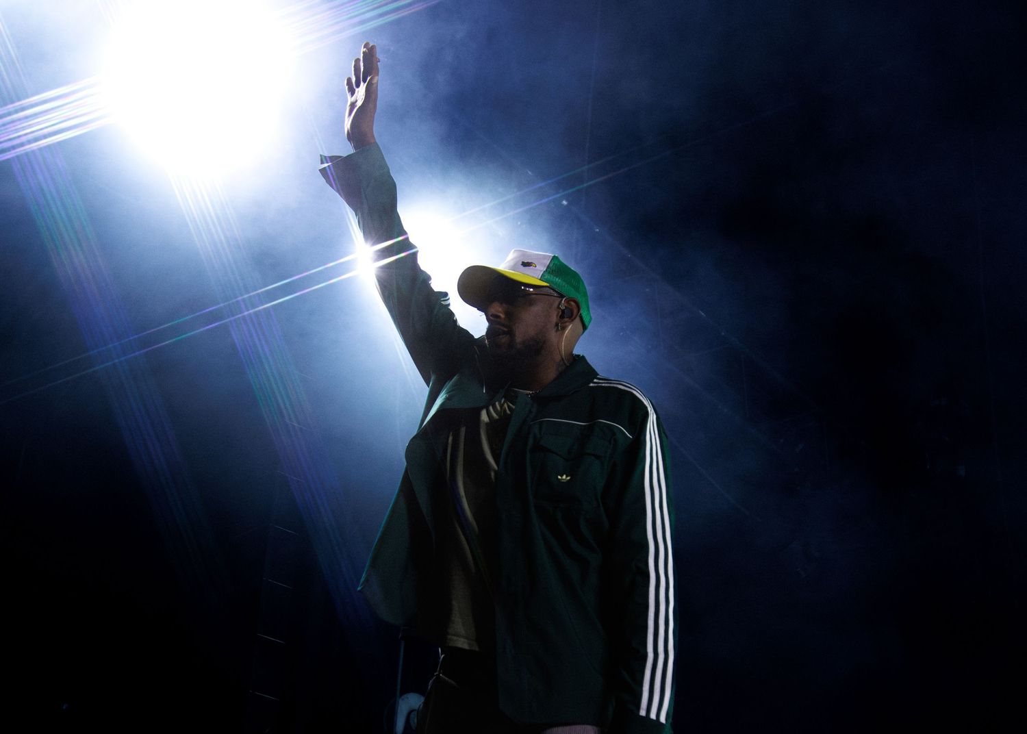 Silhouetted performer in an Adidas jacket gestures dramatically under dramatic blue stage lighting during a concert.