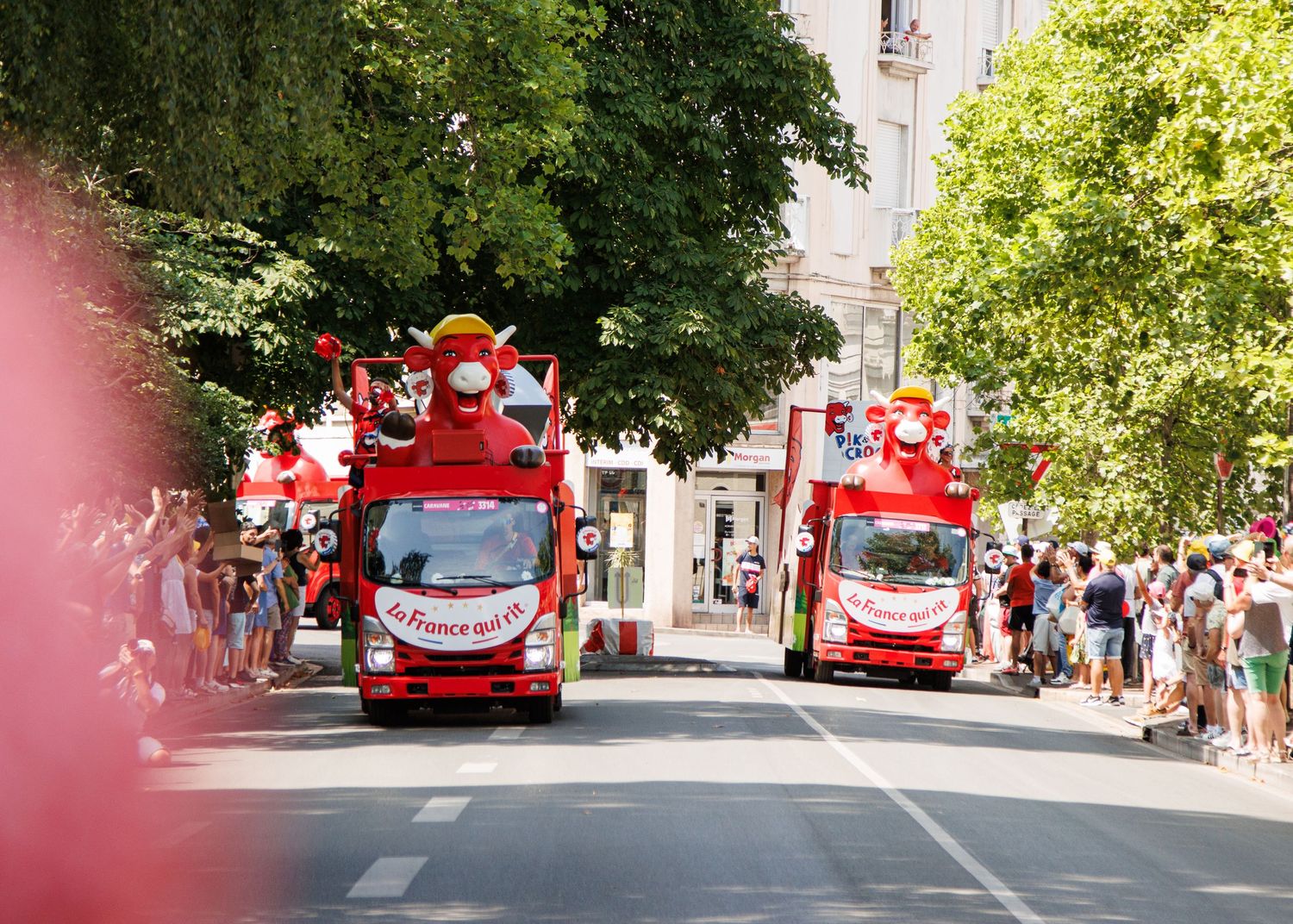 Two red promotional vehicles driving down a street lined with spectators and trees.