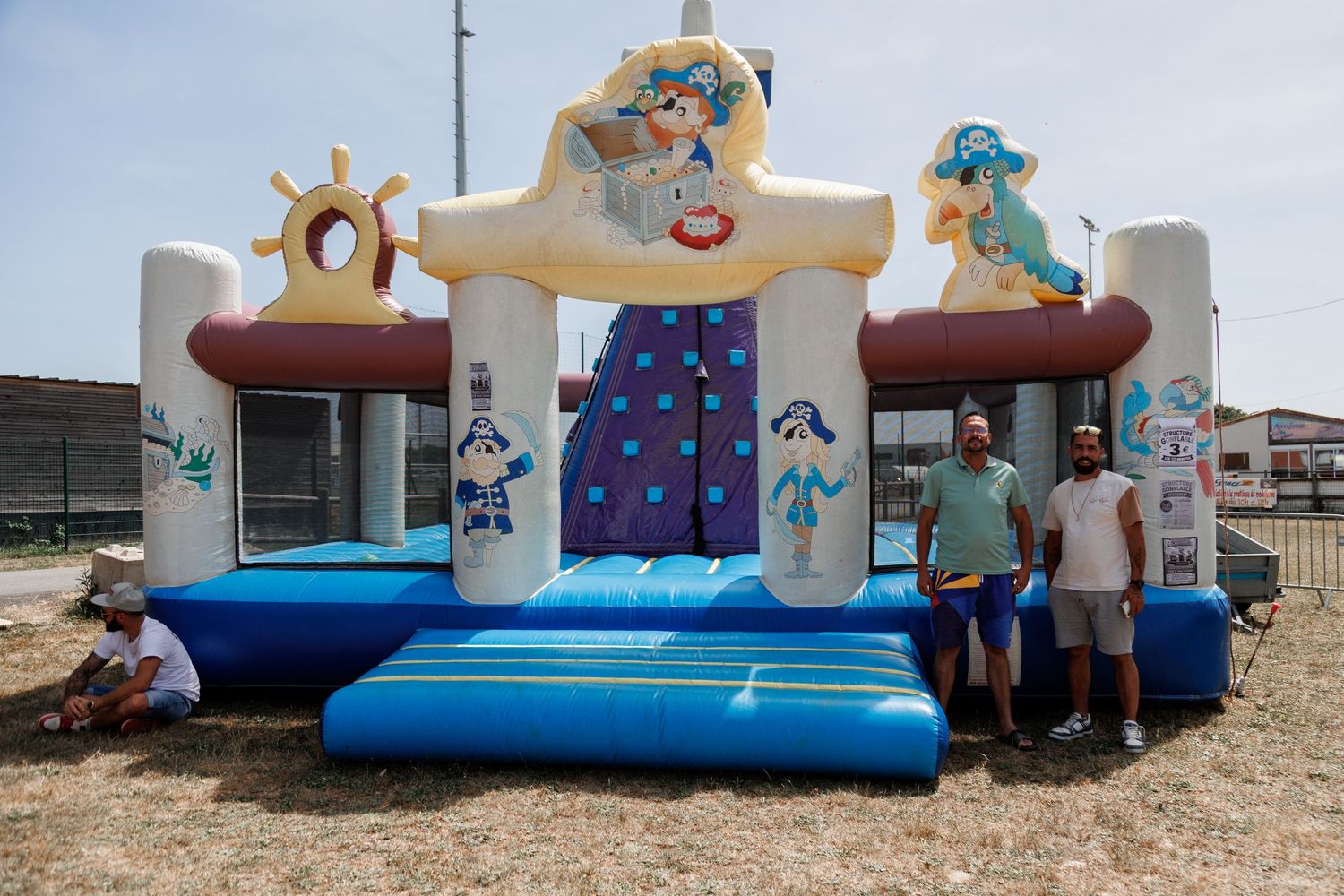 Two men stand in front of a large pirate-themed inflatable bounce house on a sunny day outdoors.