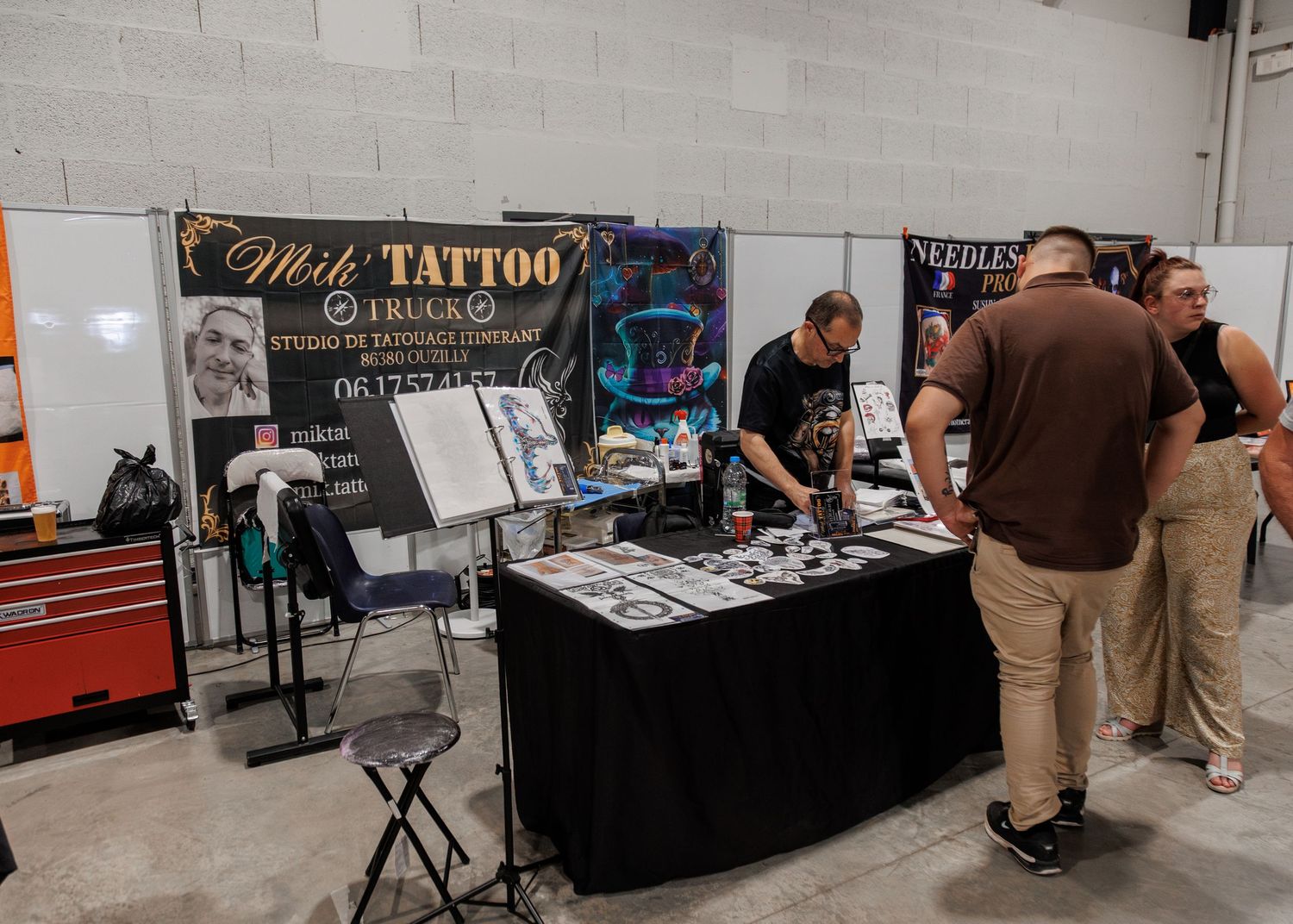 Milk Tattoo studio booth at a tattoo convention with artwork displays and visitors browsing flash designs on a black table.