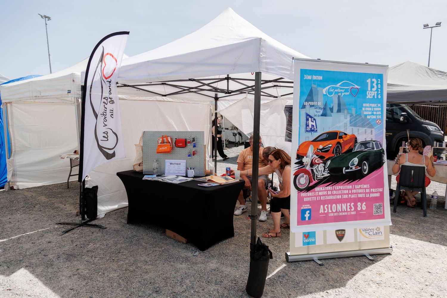 Outdoor event booth with white canopy tent, black table, and promotional banners at a sunny outdoor fair.