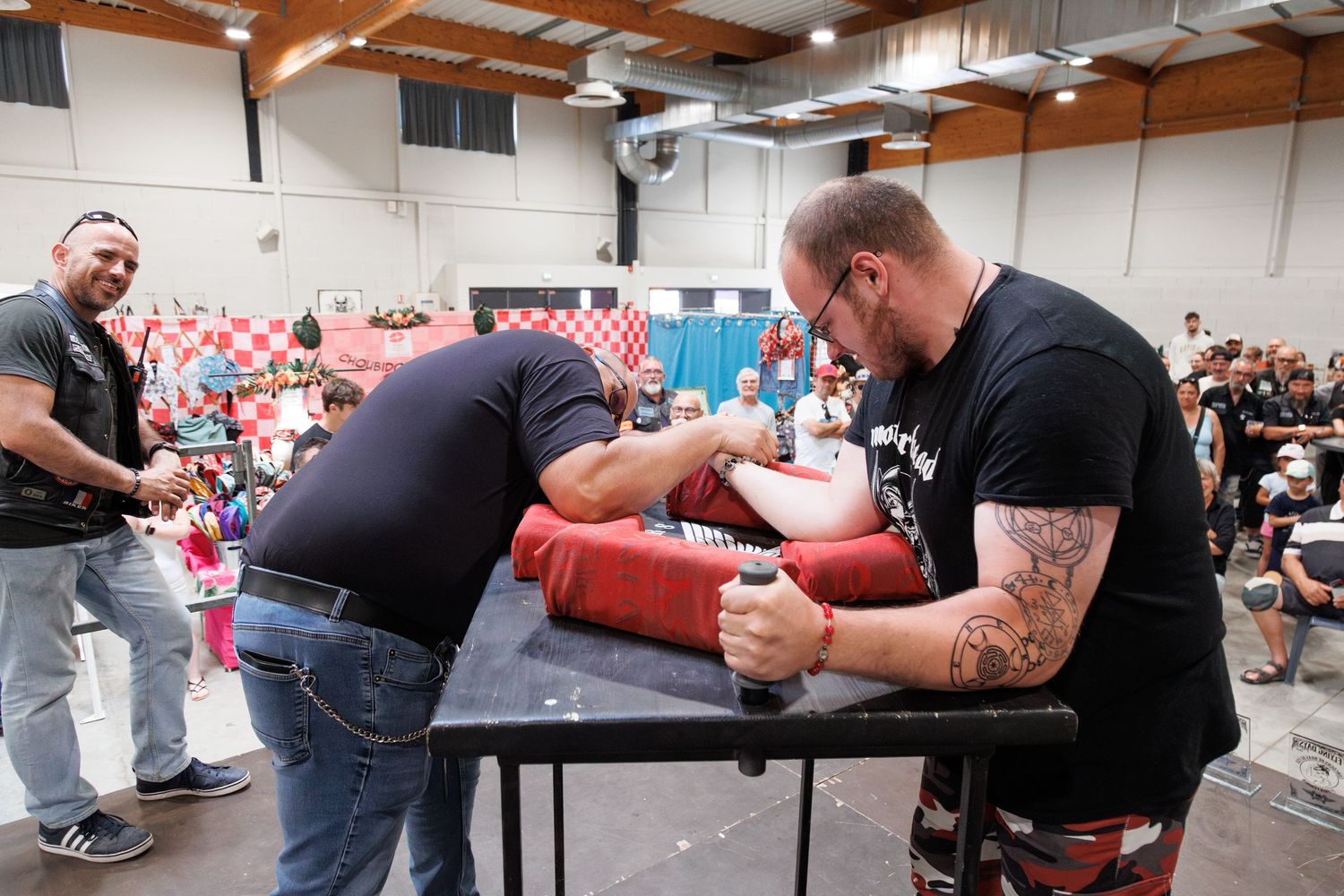 Two men competing in an arm wrestling match at an indoor tournament with spectators watching in the background.