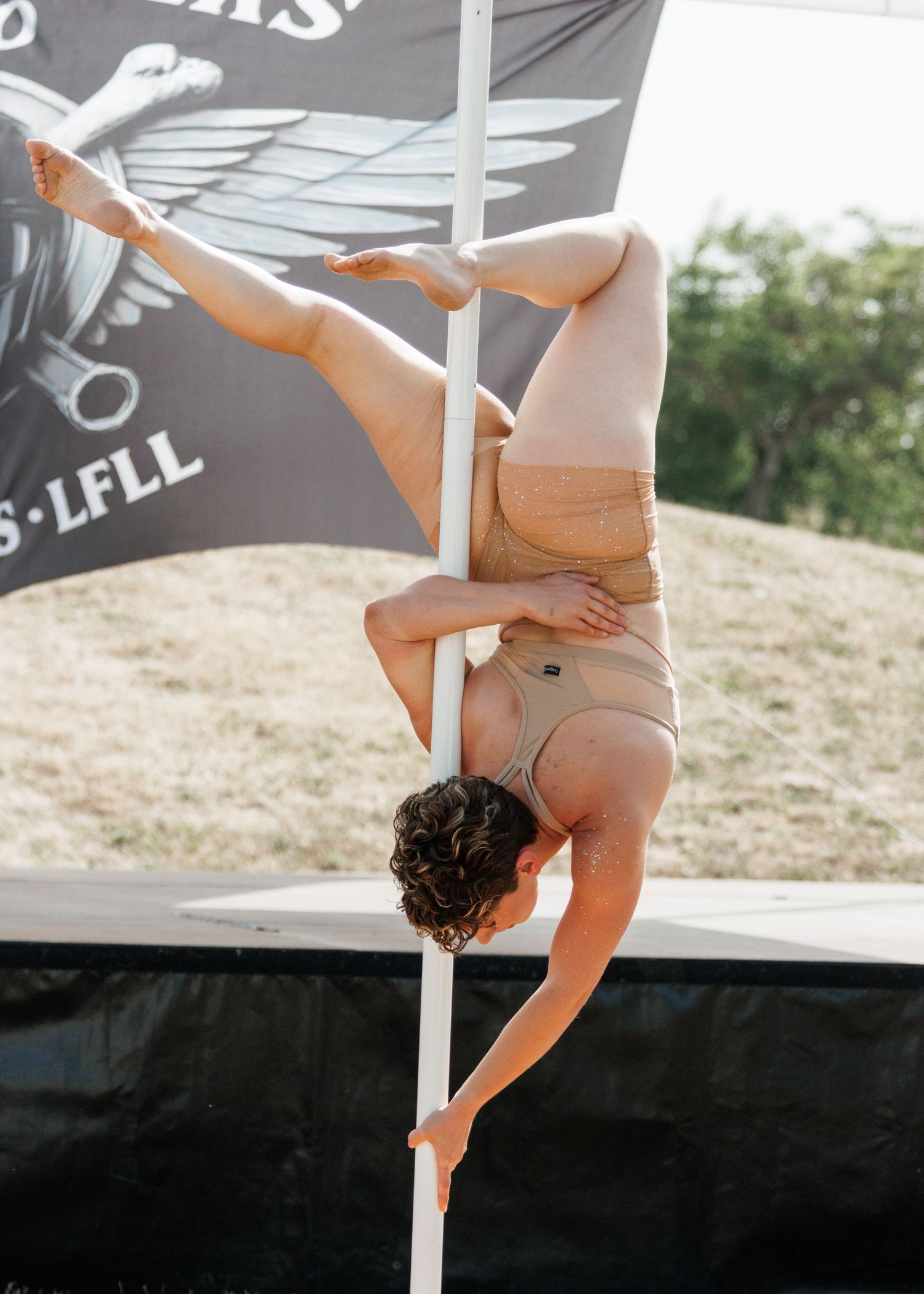 Athlete performs an inverted pole dance move outdoors in a beige outfit at an event with a dark banner in the background.