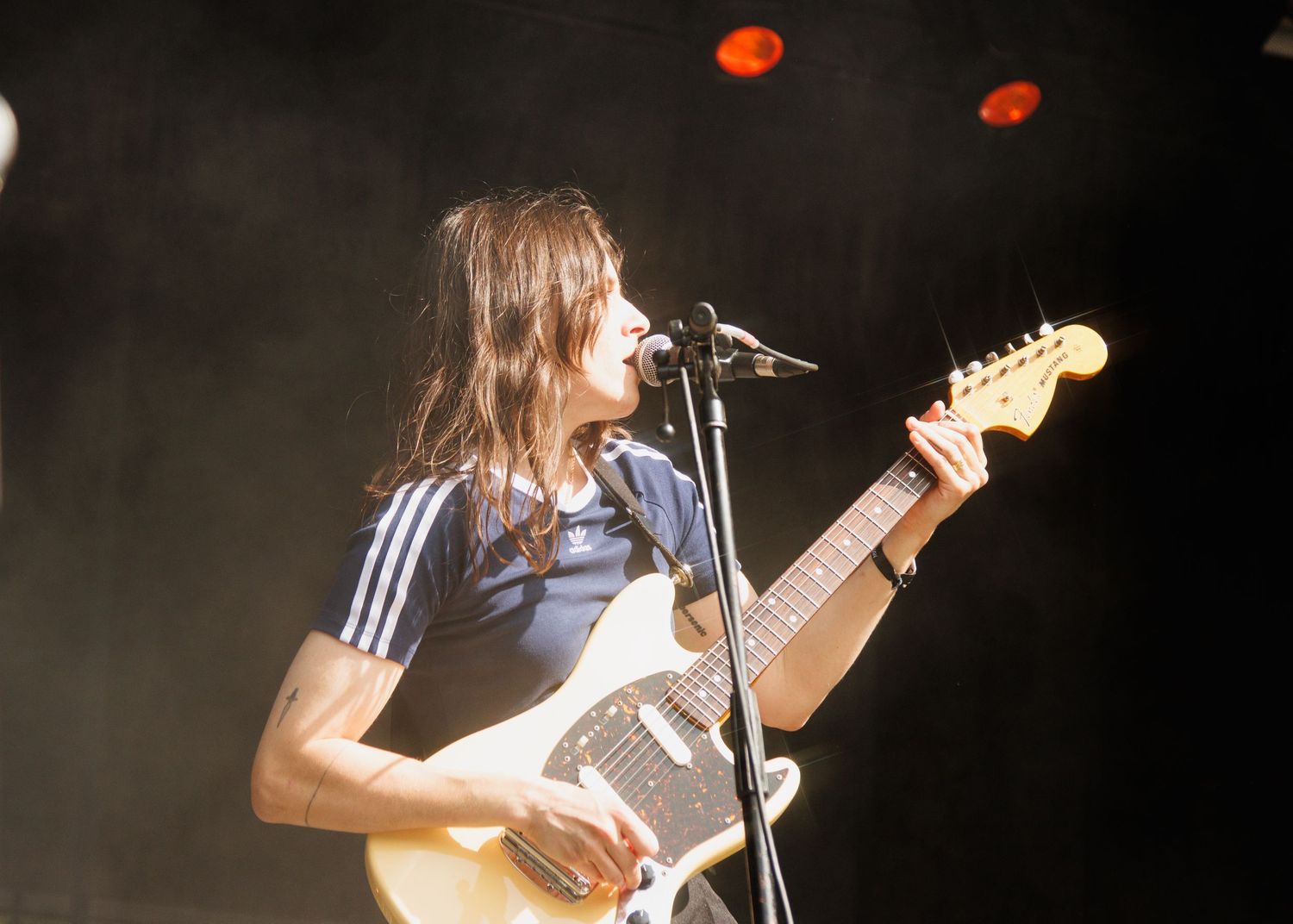 Male guitarist singing into a microphone on stage, wearing a blue striped shirt while playing a Fender guitar.