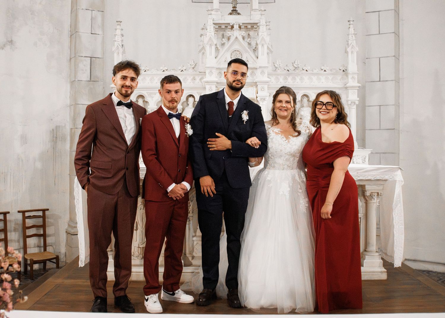 Wedding party posing in elegant white church, bride in white gown with groom and guests in burgundy formal attire.