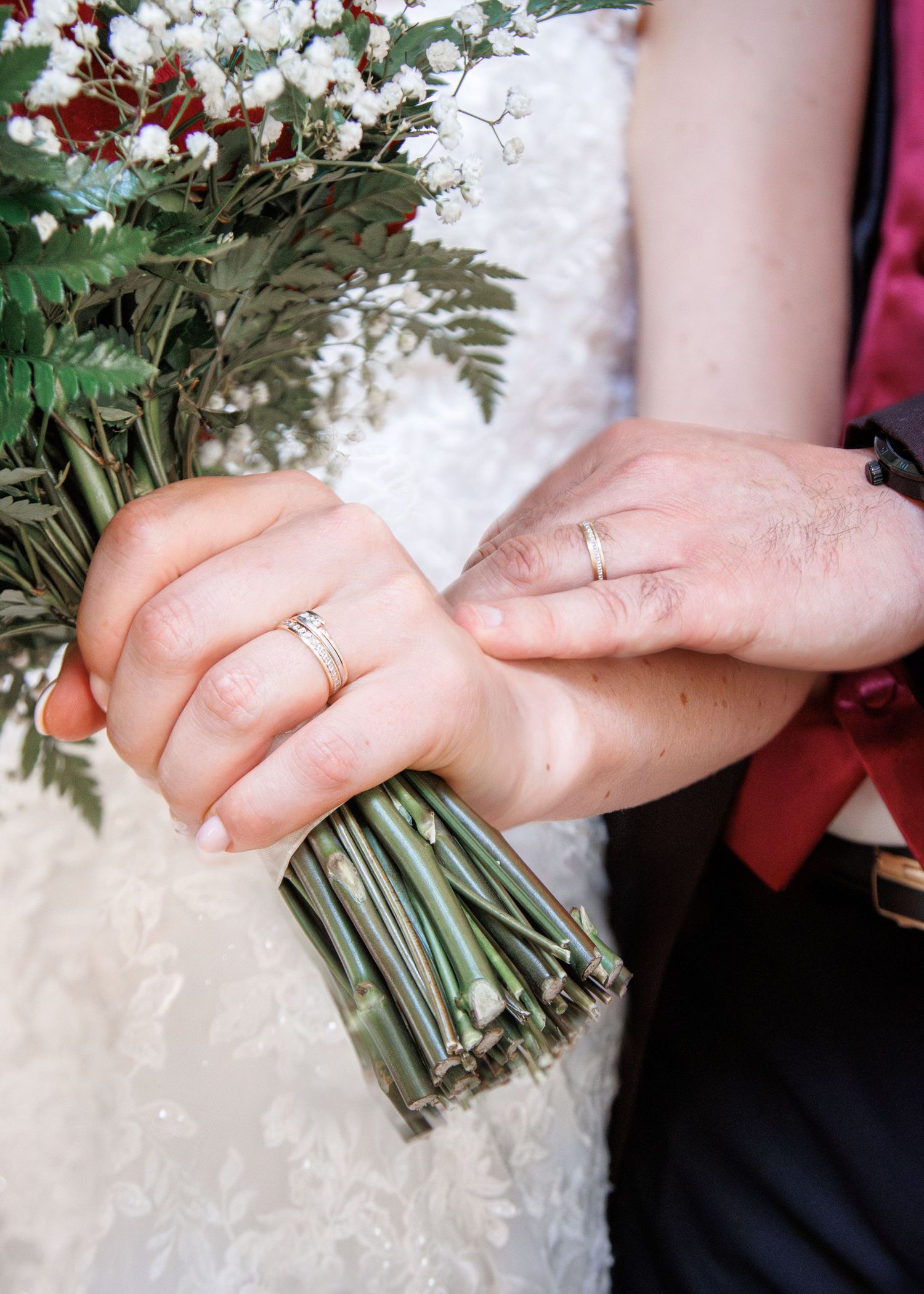 Bride and groom holding a green-stemmed wedding bouquet with white flowers, both wearing gold wedding bands.