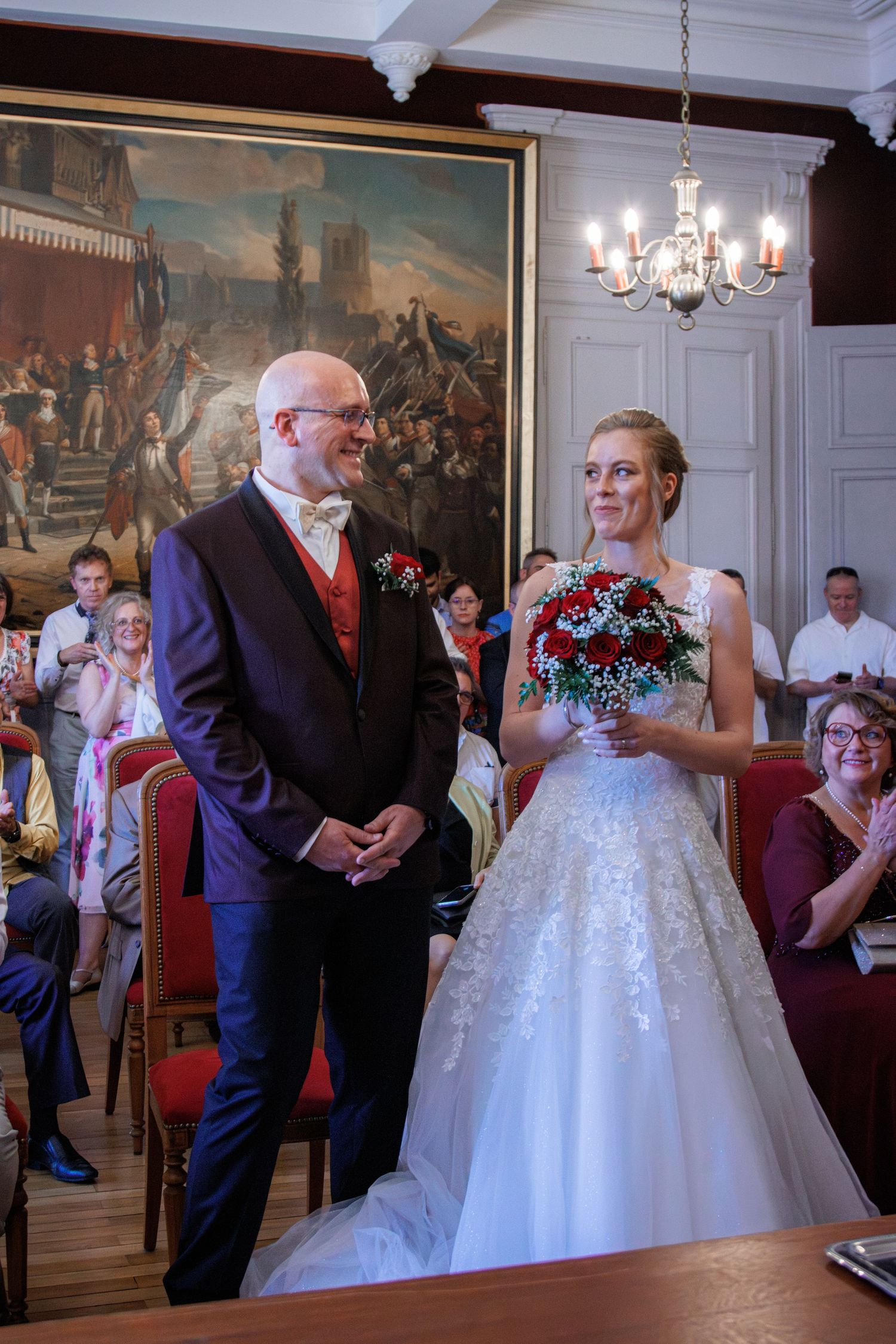 Bride in white lace gown holding red rose bouquet stands with groom in navy suit during wedding ceremony.