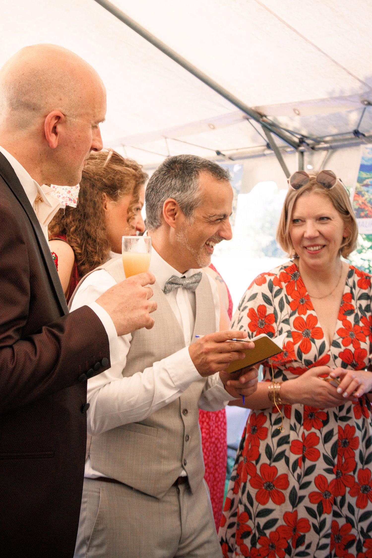 Elegantly dressed wedding guests socialize and laugh together under a tent at a daytime reception.