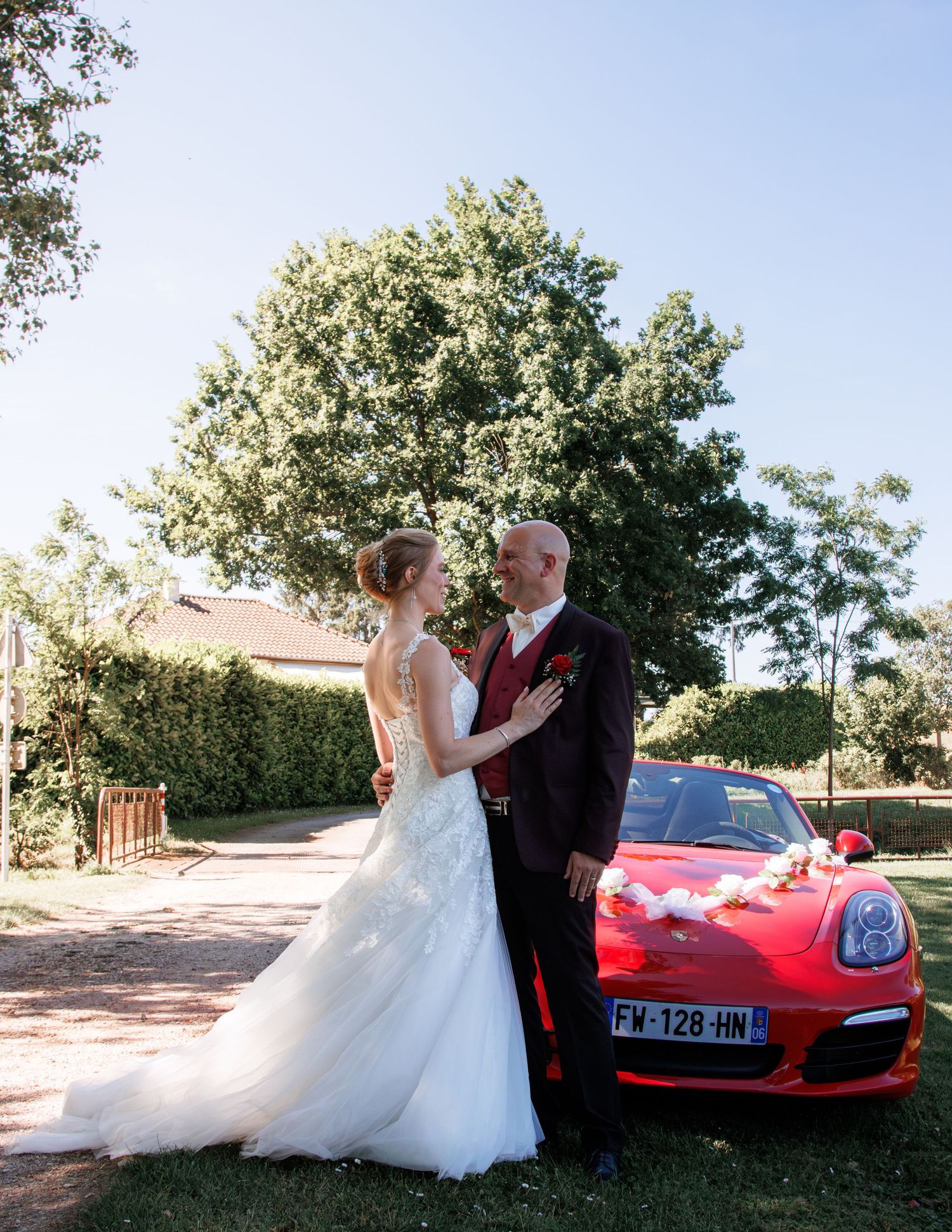 Bride and groom share romantic moment beside decorated red Porsche wedding car in sunny outdoor garden setting.