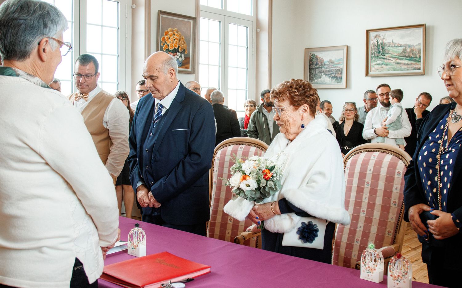 Elderly couple exchanging vows at a formal civil wedding ceremony in an elegant room with guests gathered around.