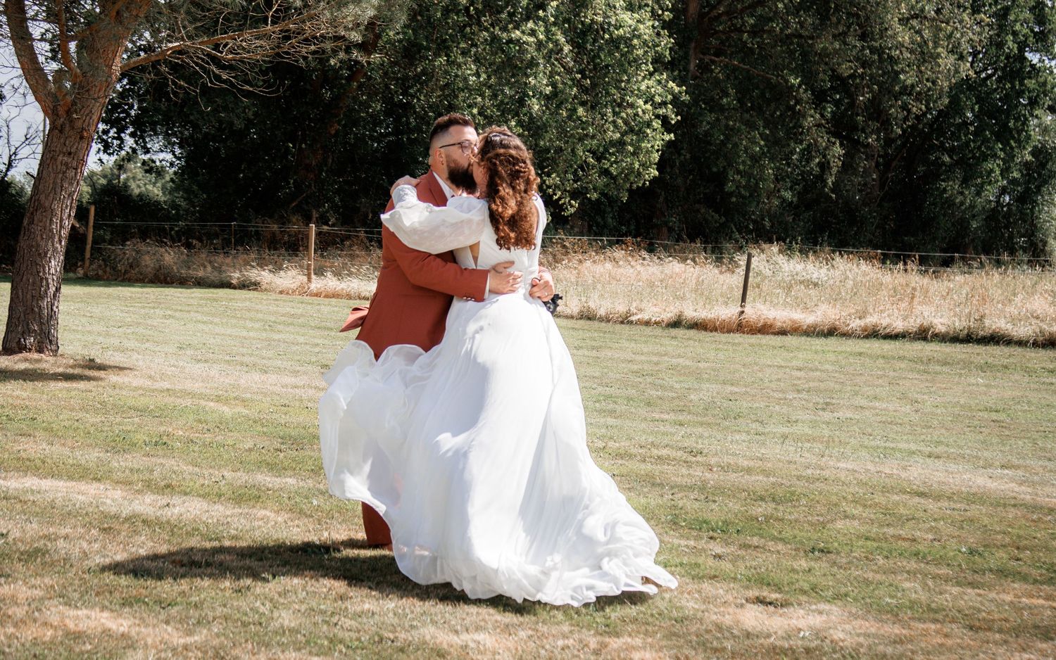 A bride in a white gown and groom in a rust suit share a romantic kiss outdoors on a sunny wedding day.