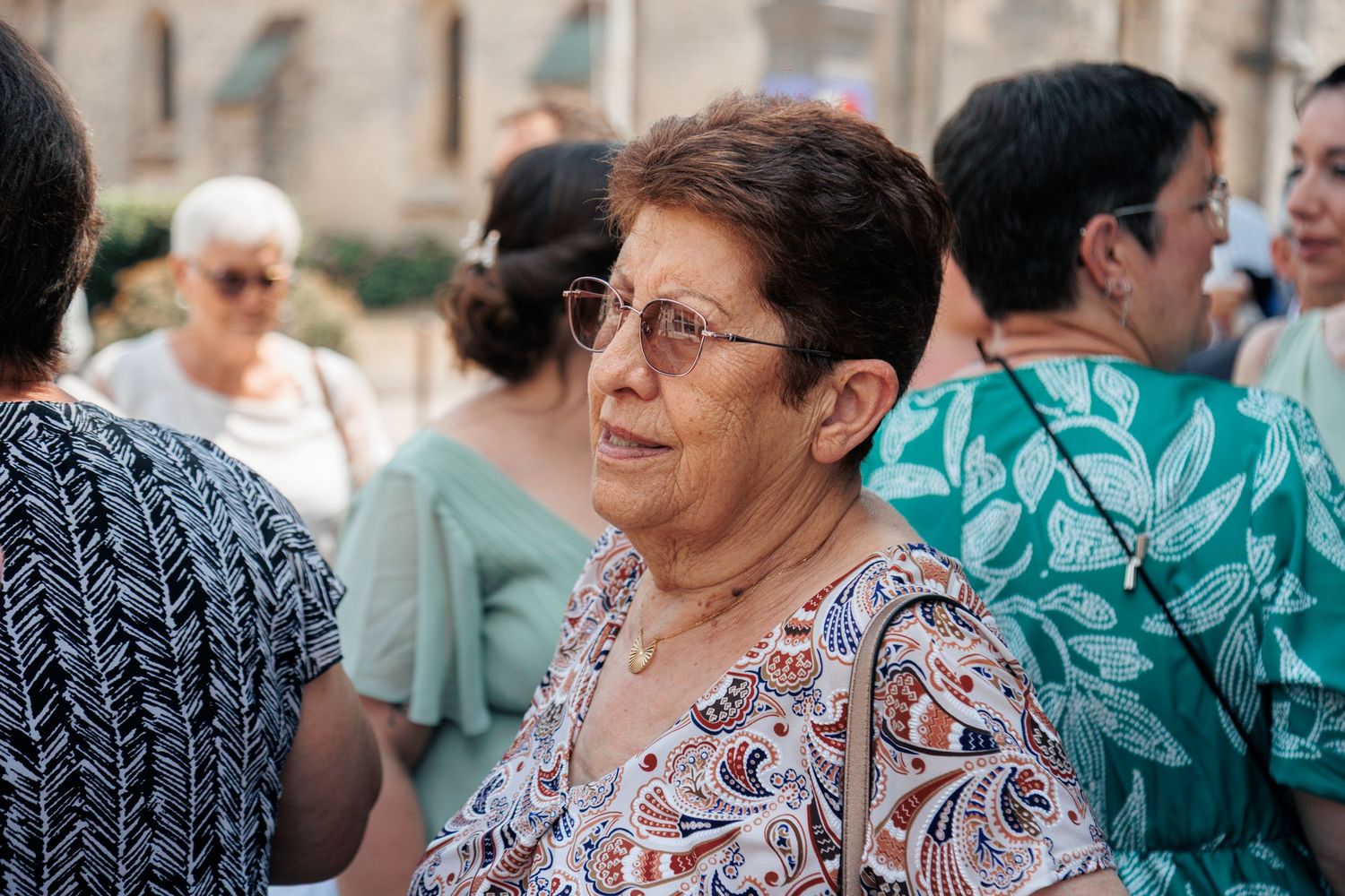 Elegant older woman with glasses and floral blouse standing among guests at an outdoor wedding gathering.