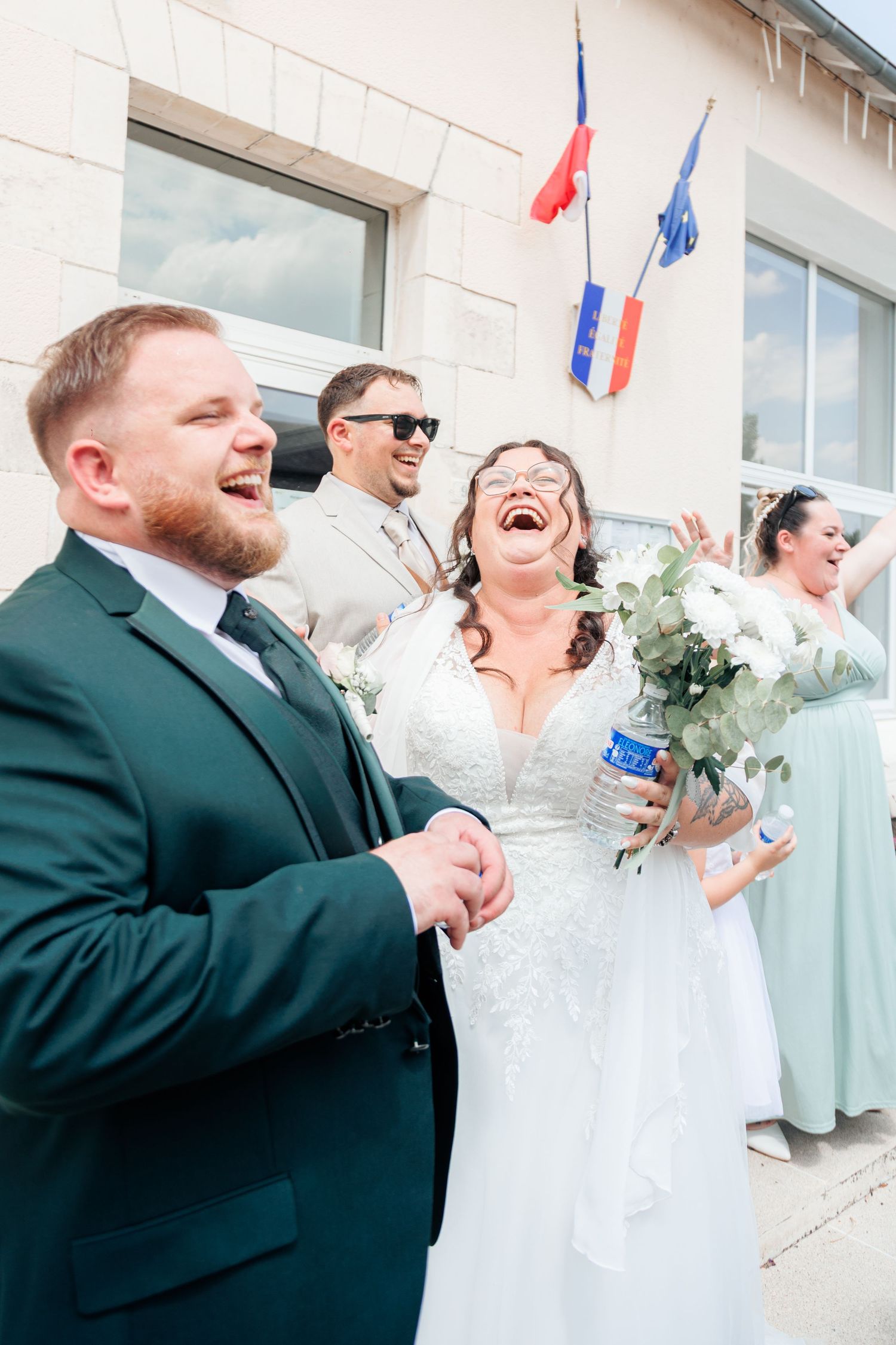 Joyful bride and groom laughing outside a building with French flags during their wedding ceremony.
