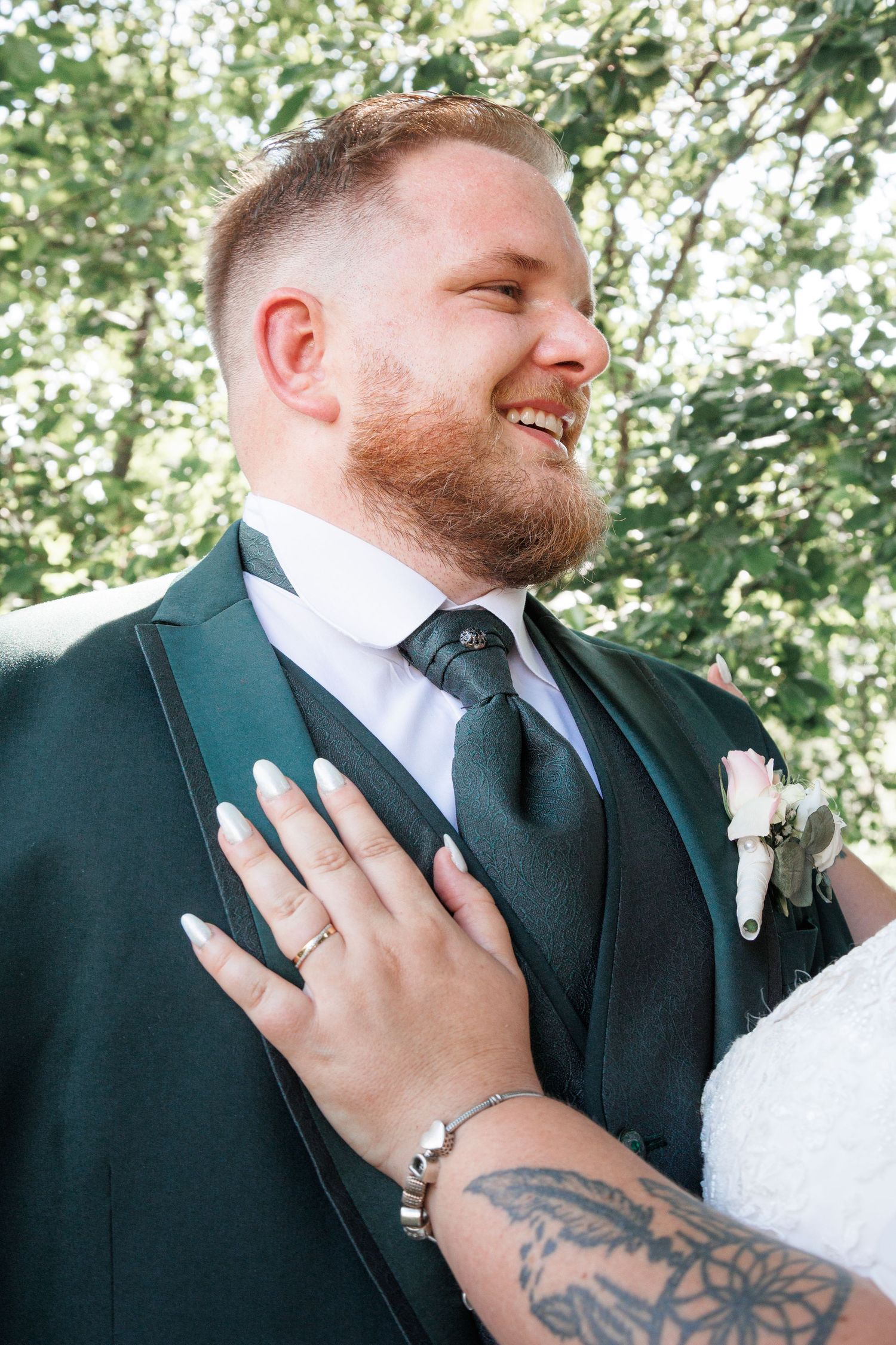 Smiling groom in dark green suit with boutonniere as bride places hand on his chest.