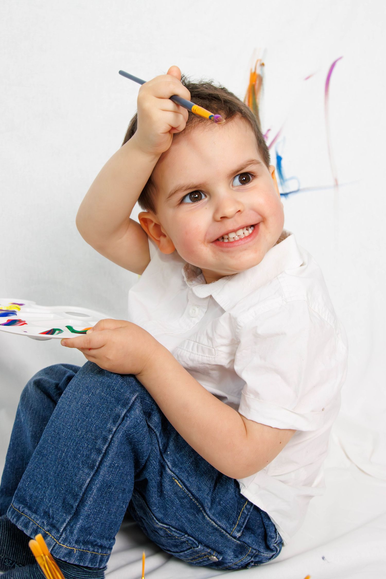Young child in white shirt and jeans smiles while holding art supplies against white background.