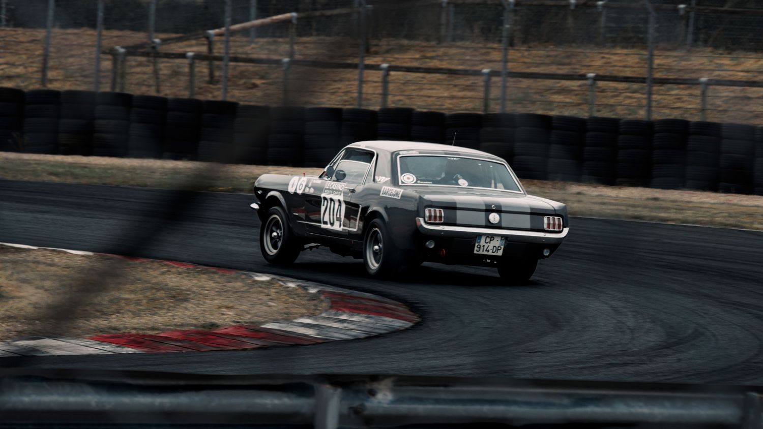 Classic sports cars compete on a race circuit with brick wall barriers visible in the background.
