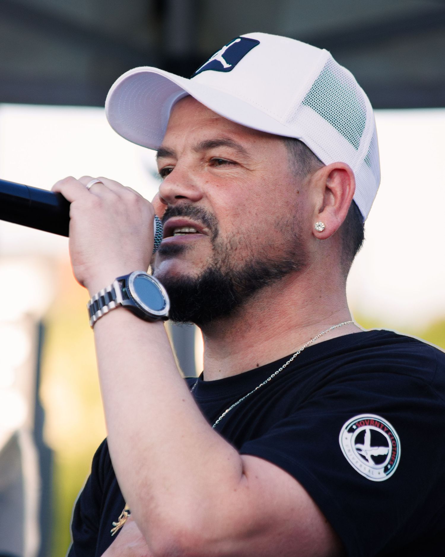 Performer in white cap and black shirt with earring holding microphone during outdoor show.