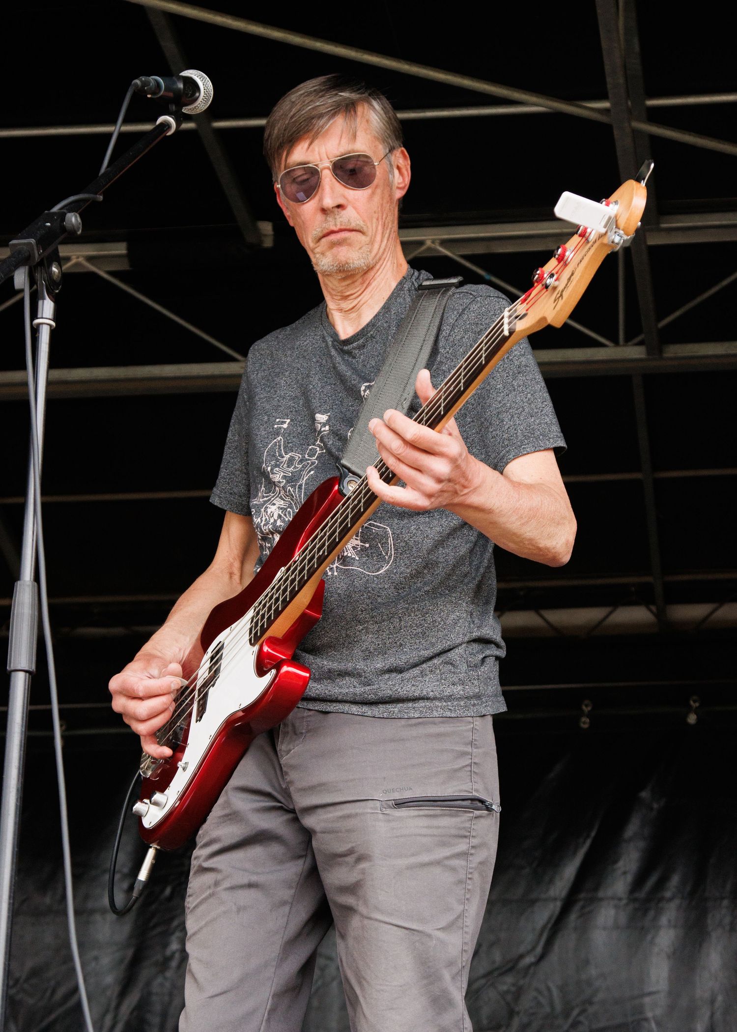 Guitarist in gray t-shirt performs on outdoor stage with red electric guitar.