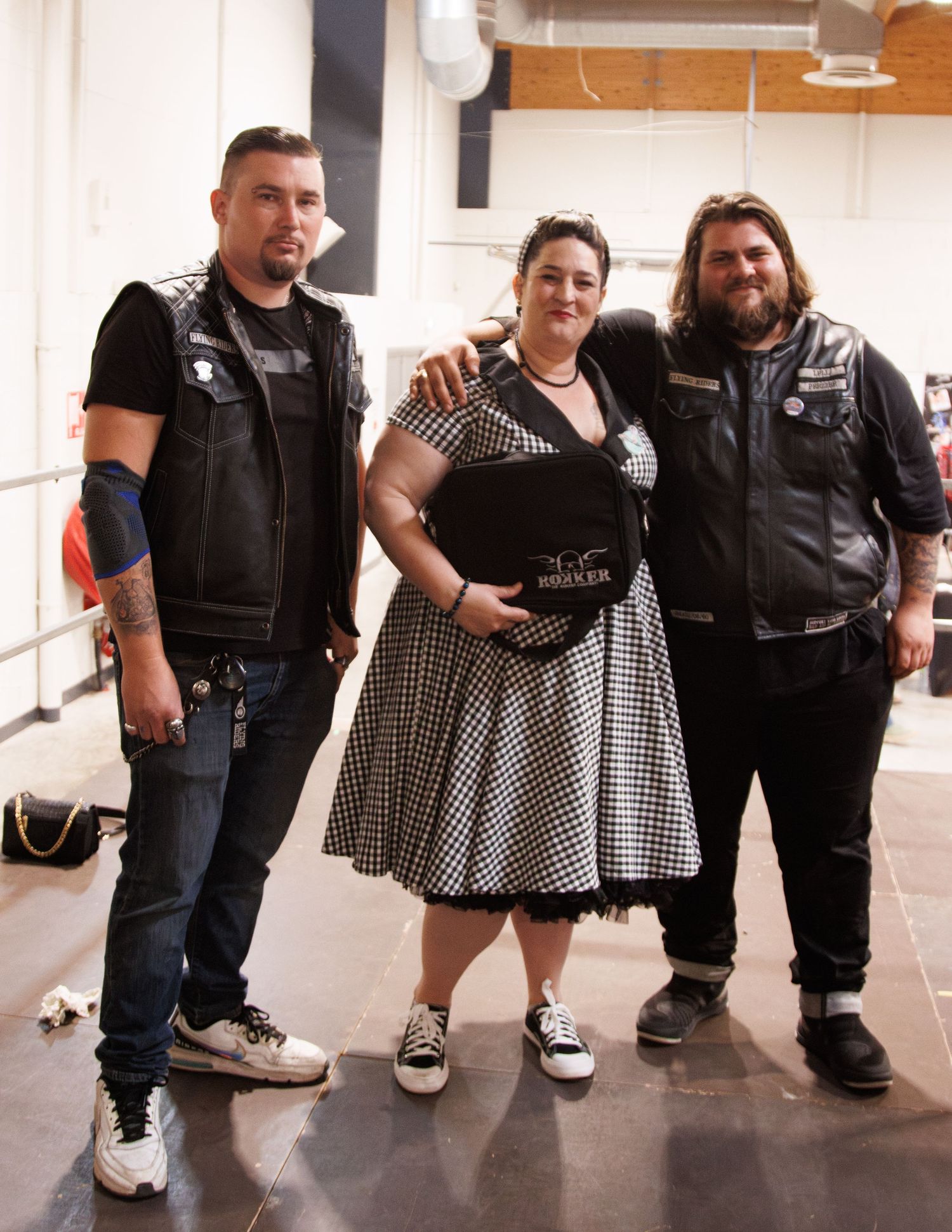Group of bikers in leather vests posing with someone in a polka dot dress for a casual outdoor gathering.